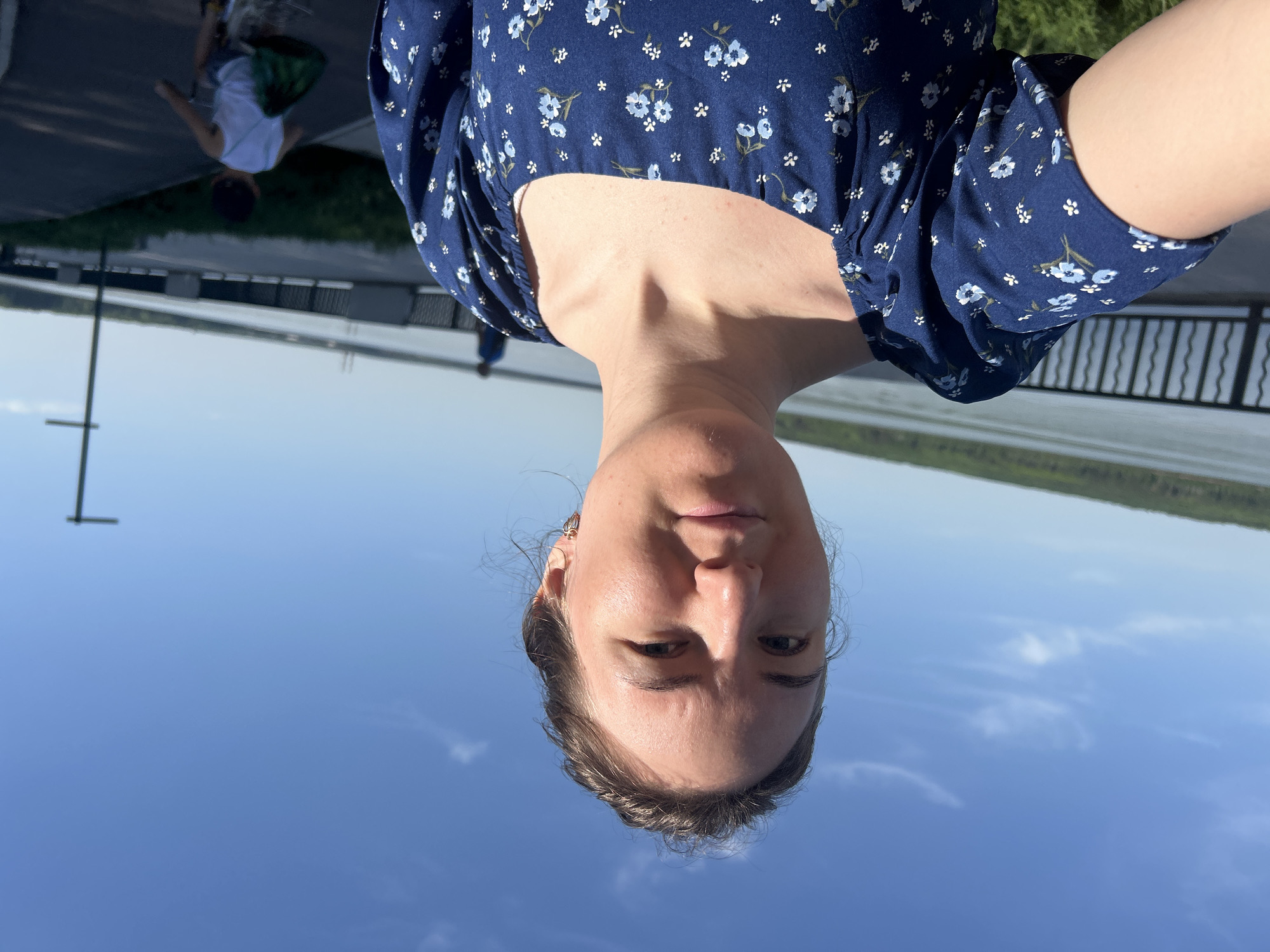 A woman in a blue floral dress is taking a selfie outdoors. The background includes a body of water and a blue sky. In the distance, a person wearing a white shirt is walking on a paved path. The photo may be taken in or near Chaikovsky, Russia.
