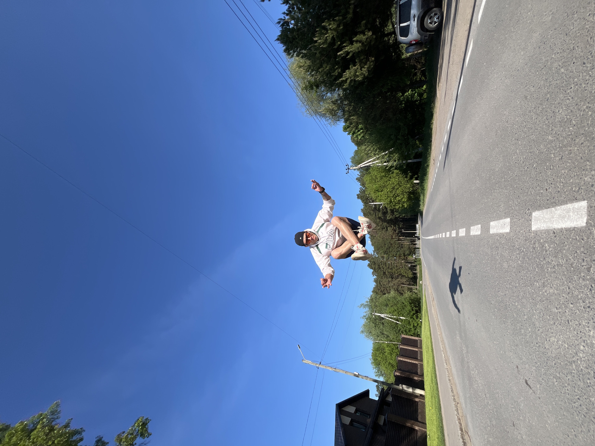 A full body shot, taken from a low-angle view, captures a person in mid-air on a paved road. The individual is wearing a white hooded sweatshirt with a "FILA" logo, dark shorts, a dark cap, and light-colored sneakers. They are gesturing with both hands, appearing to be in a squatting or crouching position.
The backdrop is a clear blue sky with utility wires stretching across. Trees line the side of the road, with a car partially visible on the right. A shadow of the person is cast onto the road surface. The road has white lane markings and is situated near a building on the left side.
The location is likely Chaikovsky, Russia, based on the surrounding buildings and foliage.