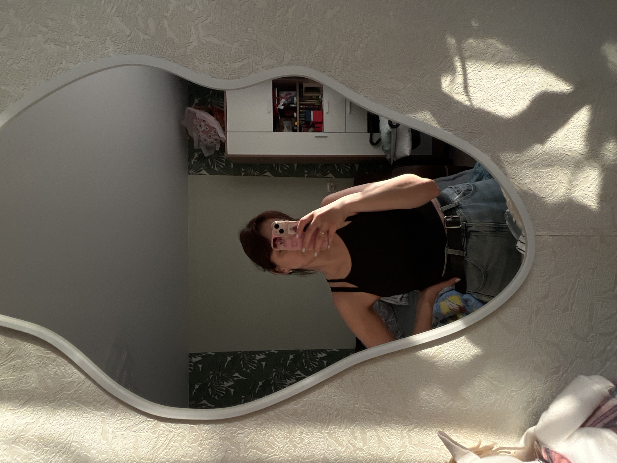 A woman is taking a selfie in a uniquely shaped mirror. The mirror reflects a room with white walls decorated with patterned wallpaper. A white cabinet with a shelf of belongings sits against the wall. The woman is wearing a black tank top and jeans with a black belt. The location is likely a private residence, but its location within the city of Saint Petersburg, Russia, cannot be determined from the visual data.