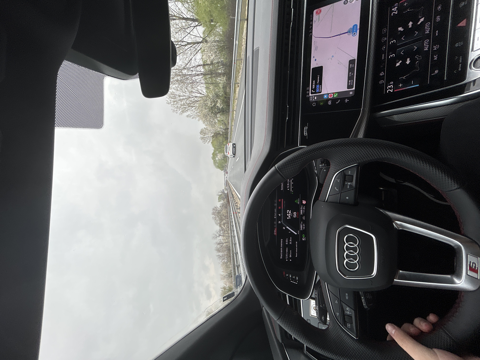 The image shows the interior of an Audi car, specifically the driver's perspective, while traveling on a highway. The speedometer reads 142 km/h. The navigation system is displaying a route approximately 108 km away, suggesting a destination involving Pernik and Kulata. The location is near Malo Konare, Bulgaria. Trees line the side of the road. A hand is visible on the steering wheel. The sky is overcast.