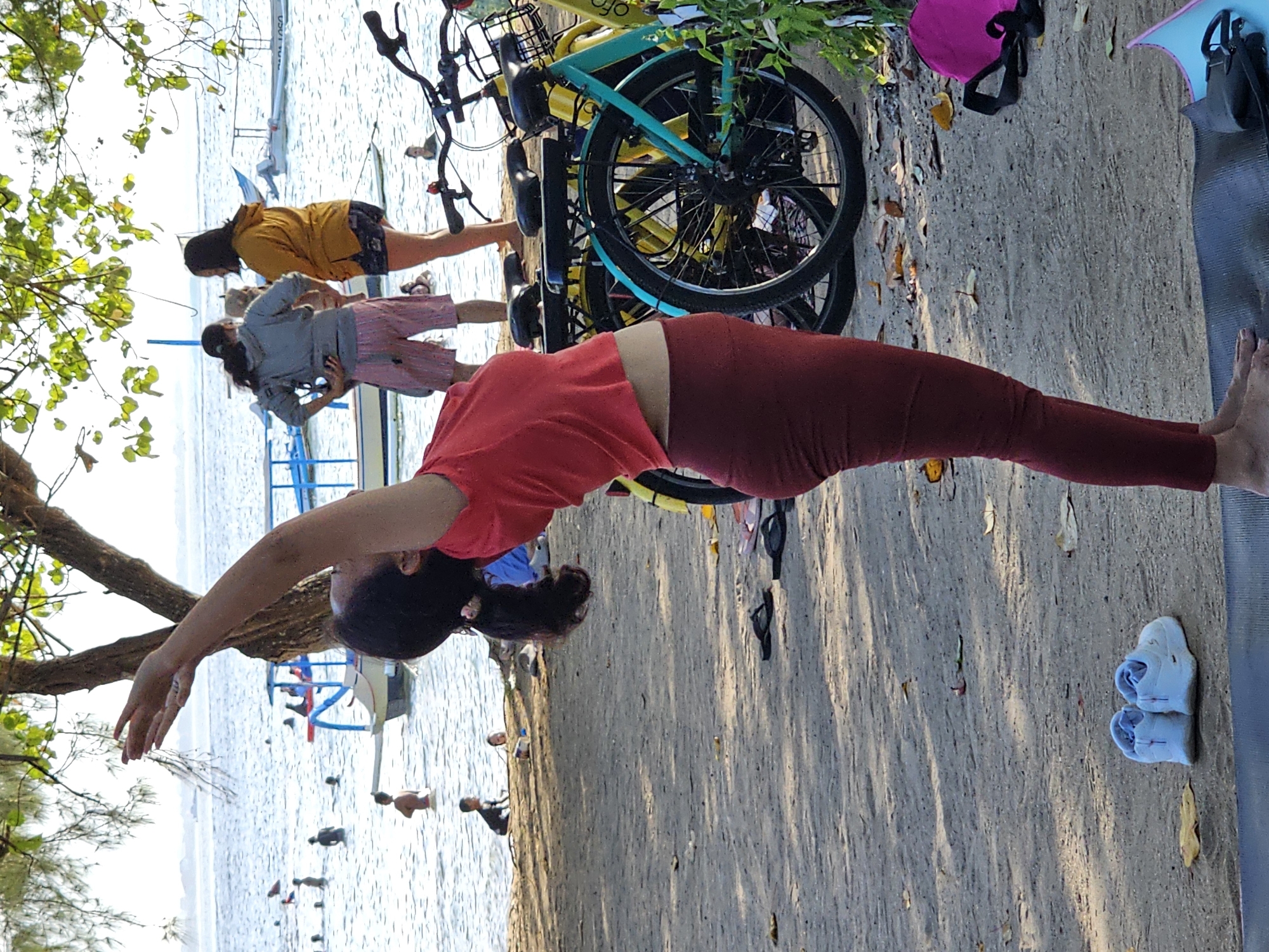 A woman in a red top and maroon leggings performs a yoga pose on a mat on a sandy beach in Denpasar, Indonesia. Her arms are raised overhead, and her body is arched backward. In the background, the ocean is visible with several people swimming and boats docked. Two bicycles, one teal and one yellow, are parked nearby, and other beachgoers are in the distance. The scene appears to be at a public beach.