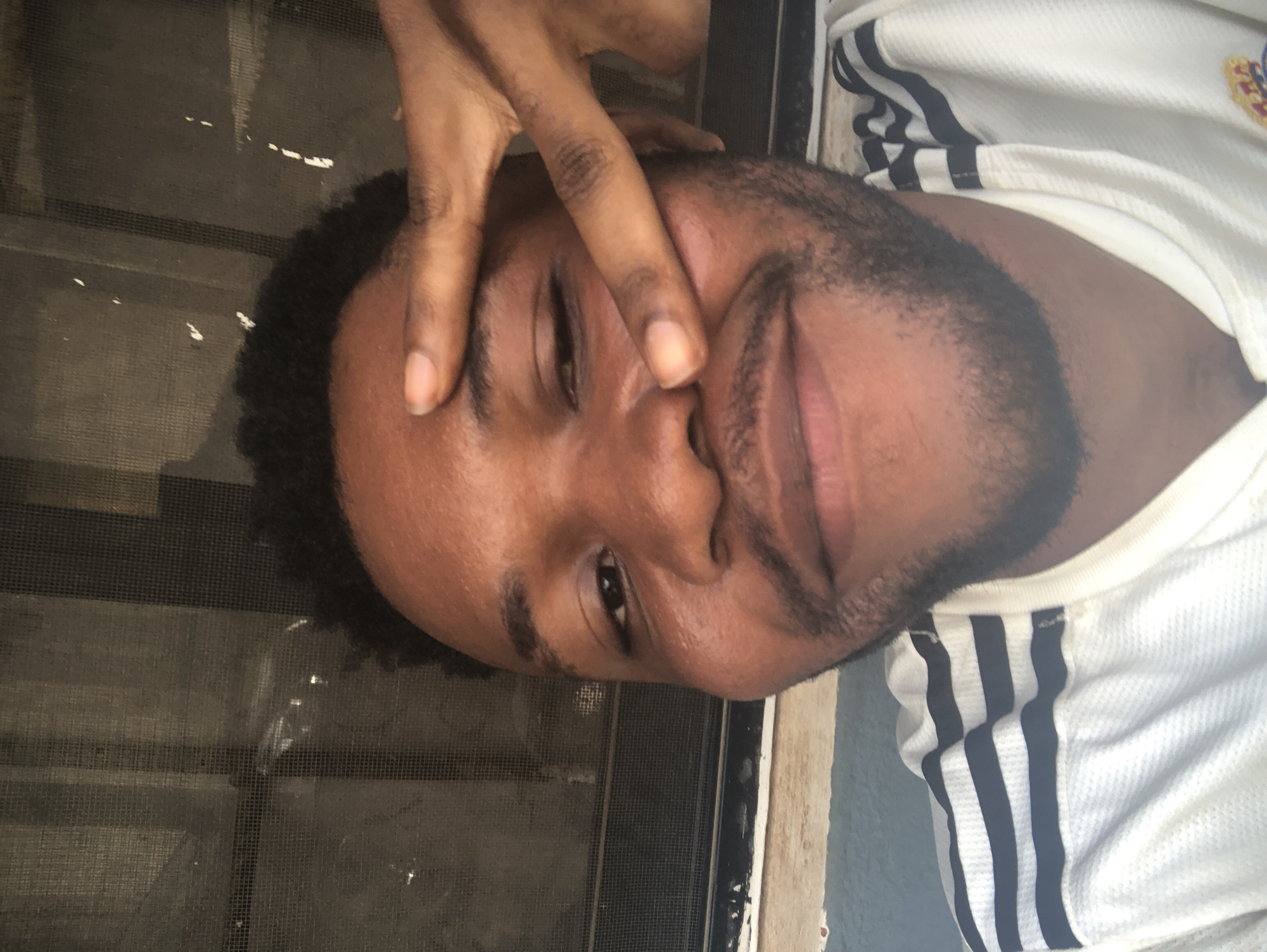 A man with dark hair and a beard is looking at the camera with a slightly smirking expression. He has his fingers on his forehead and cheek. The man is wearing a white shirt with black stripes on the sleeves. The background includes a screen. The location is Unknown, Unknown.