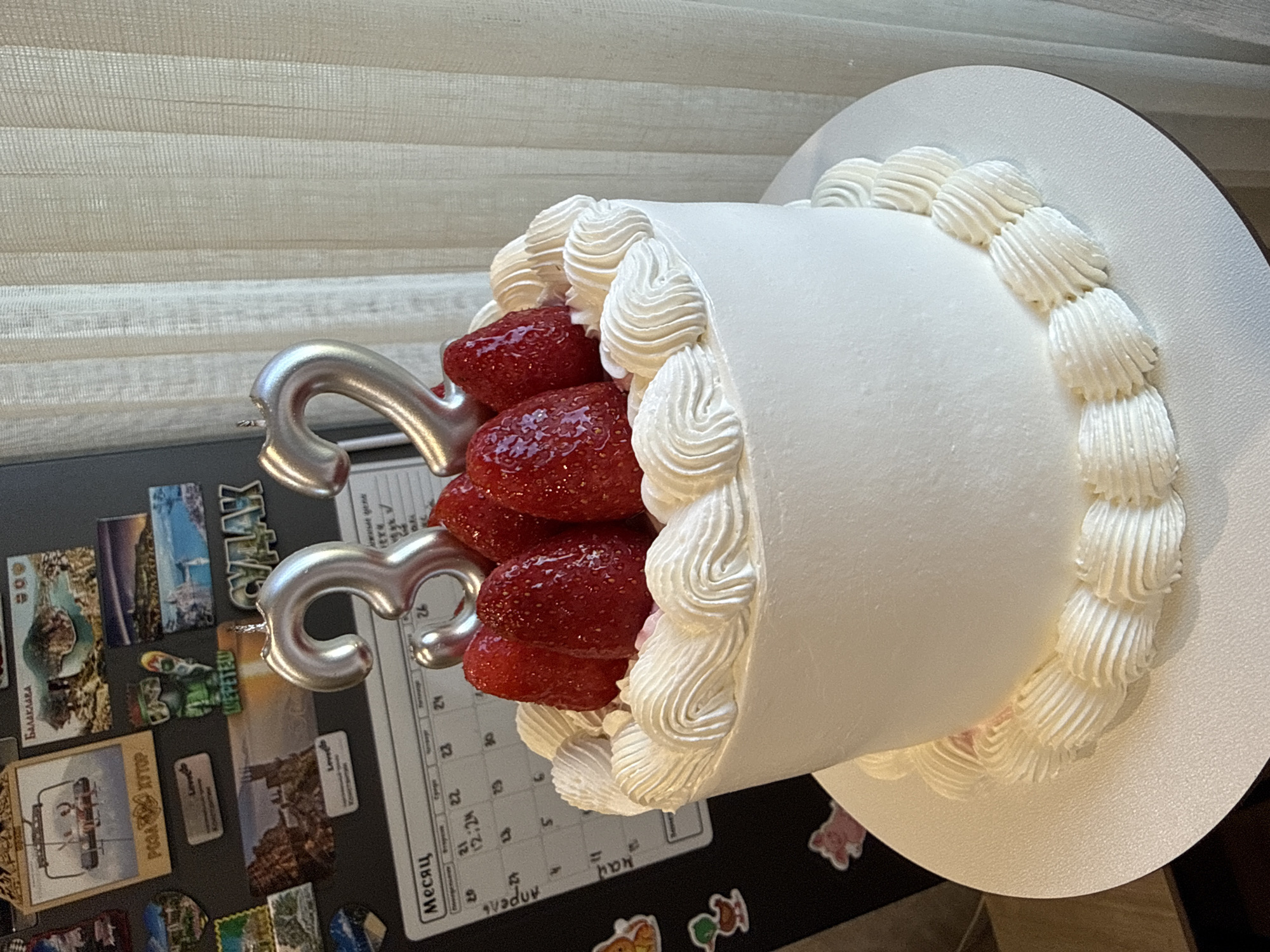 The image shows a cake decorated with strawberries and white frosting. The cake is adorned with two silver number candles, appearing to represent the number 33. The cake rests on a refrigerator door covered with various magnets and a calendar. The calendar is in Russian and lists the months of April and May, with several dates highlighted. Some of the magnets are postcards from various locations, including "РОЗА ХУТОР" and "Loved", while others are small images of animals. The background features a partially visible window covered with blinds. The overall setting suggests a domestic interior.