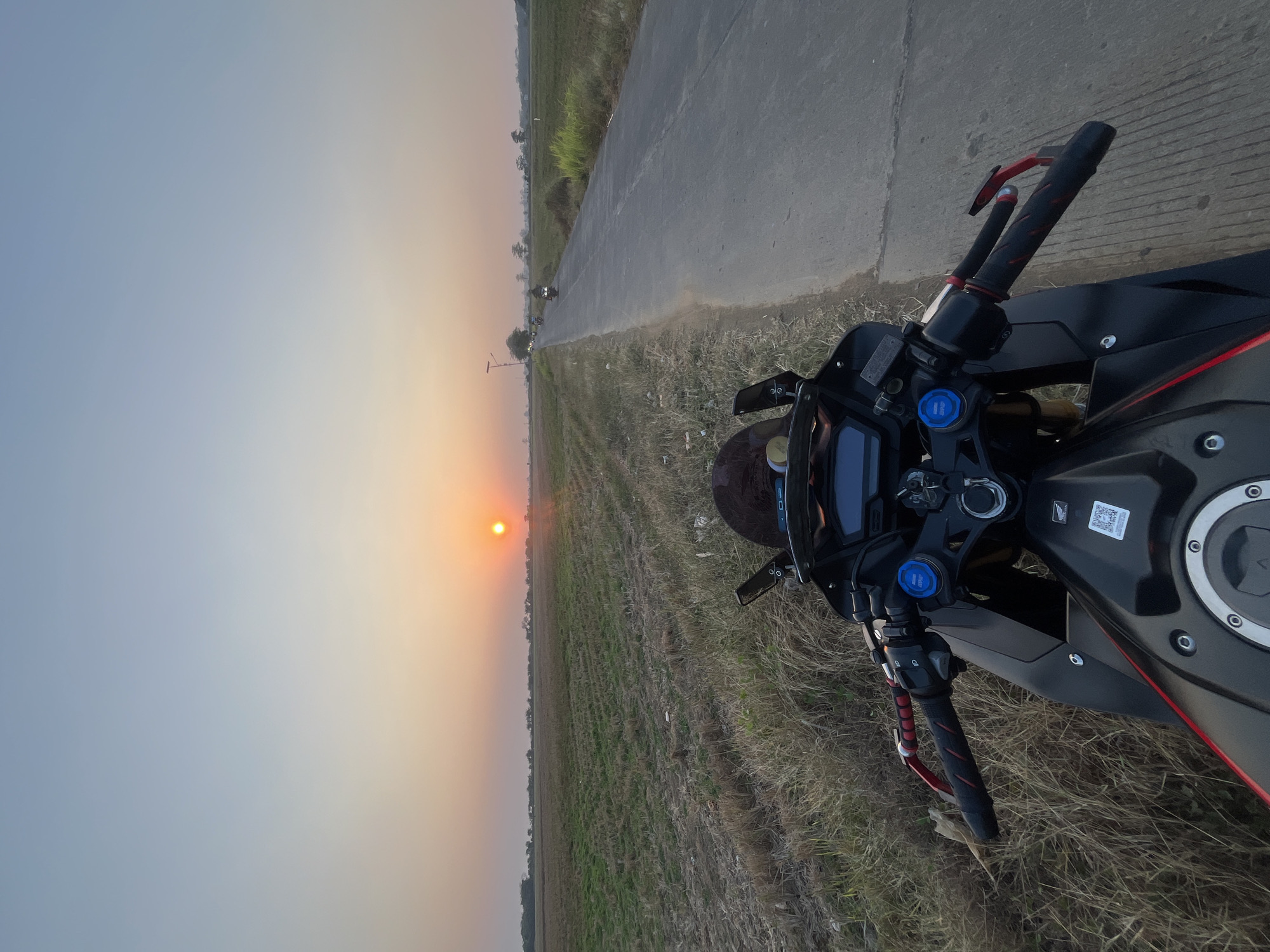 A first-person perspective shot shows the handlebars of a black sport motorcycle on the side of a paved road in a rural area. The road stretches into the distance, flanked by dry, grassy fields. In the background, the sun is setting over a flat horizon, casting a warm orange glow. Several other motorcycles are visible in the distance on the road, also traveling away from the camera. The sky transitions from a pale blue at the top to a soft orange and yellow near the horizon. The scene appears to be in a rural area of Troso, Indonesia, given the agricultural landscape and the presence of multiple motorcycles.