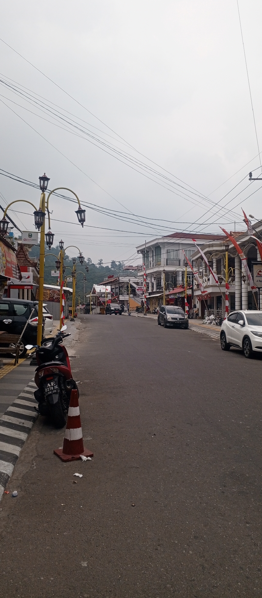 The image shows a street scene in Sarangan, Indonesia, characterized by a cloudy sky and overhead power lines. The street is paved with asphalt and has a striped pedestrian crossing on the left side. A red and white traffic cone is placed on the side of the road, next to a red motorcycle. Several vehicles, including cars and a motorcycle, are parked along the street or in motion. Buildings with traditional Indonesian flags and banners line both sides of the street. The architecture of the buildings suggests a commercial area with shops and businesses. In the background, a lush, green hillside is visible, indicating a mountainous or hilly terrain. The overall impression is that of a quiet street in a town, possibly a tourist destination, with a mix of local life and commercial activity.