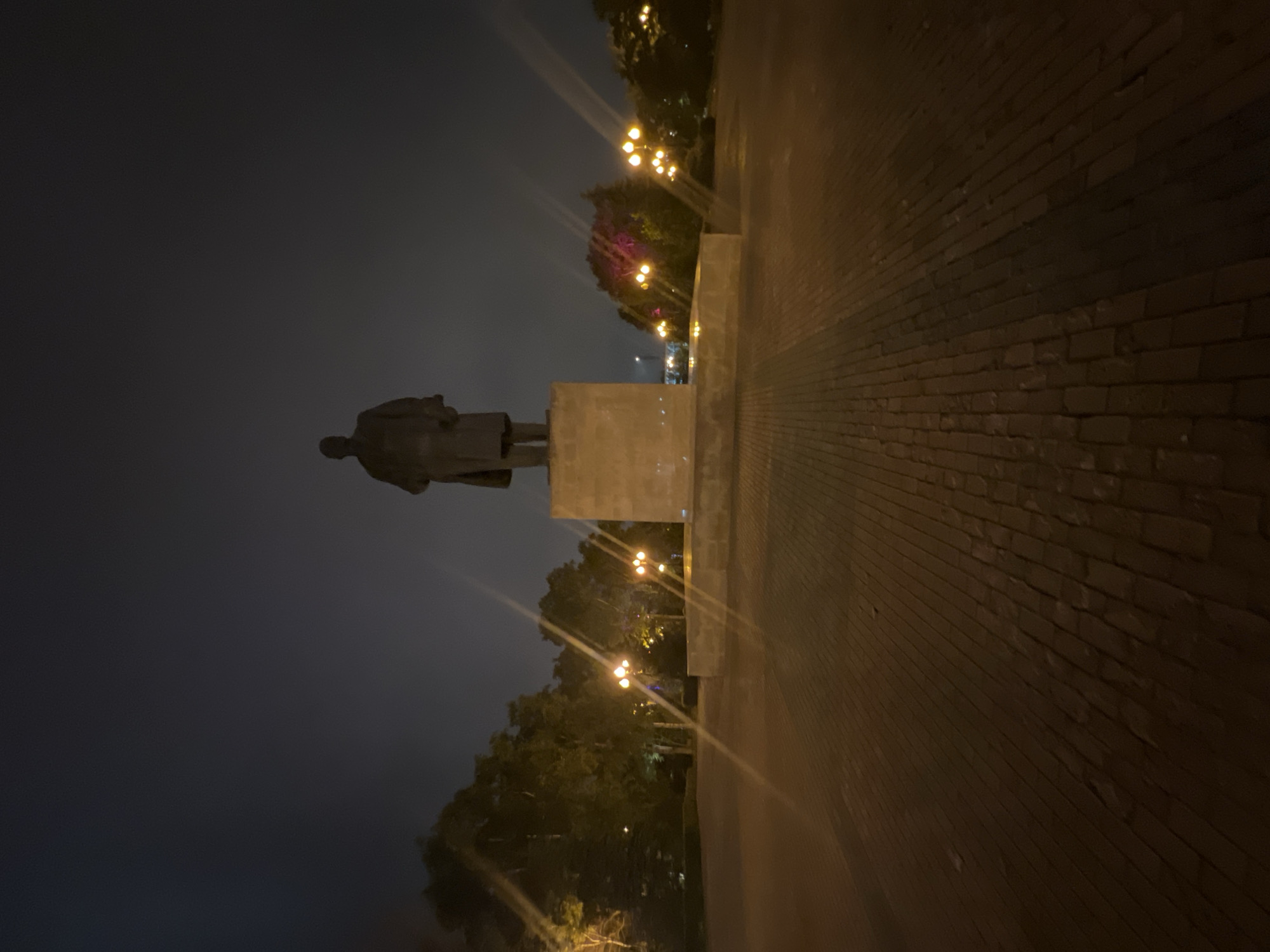The image captures a nighttime scene in Yuzhno-Sakhalinsk, Russia, featuring a prominent statue of Vladimir Lenin. The statue stands tall on a stone pedestal, silhouetted against a dark, cloudy sky. The surrounding area is a wide, paved plaza, illuminated by scattered streetlights that cast long shadows across the brickwork. Trees with foliage are visible on the left and right sides of the plaza, with some trees appearing to have colored lights, possibly purple, emanating from within them. The overall atmosphere is somber and slightly eerie due to the low light conditions and the imposing presence of the statue.