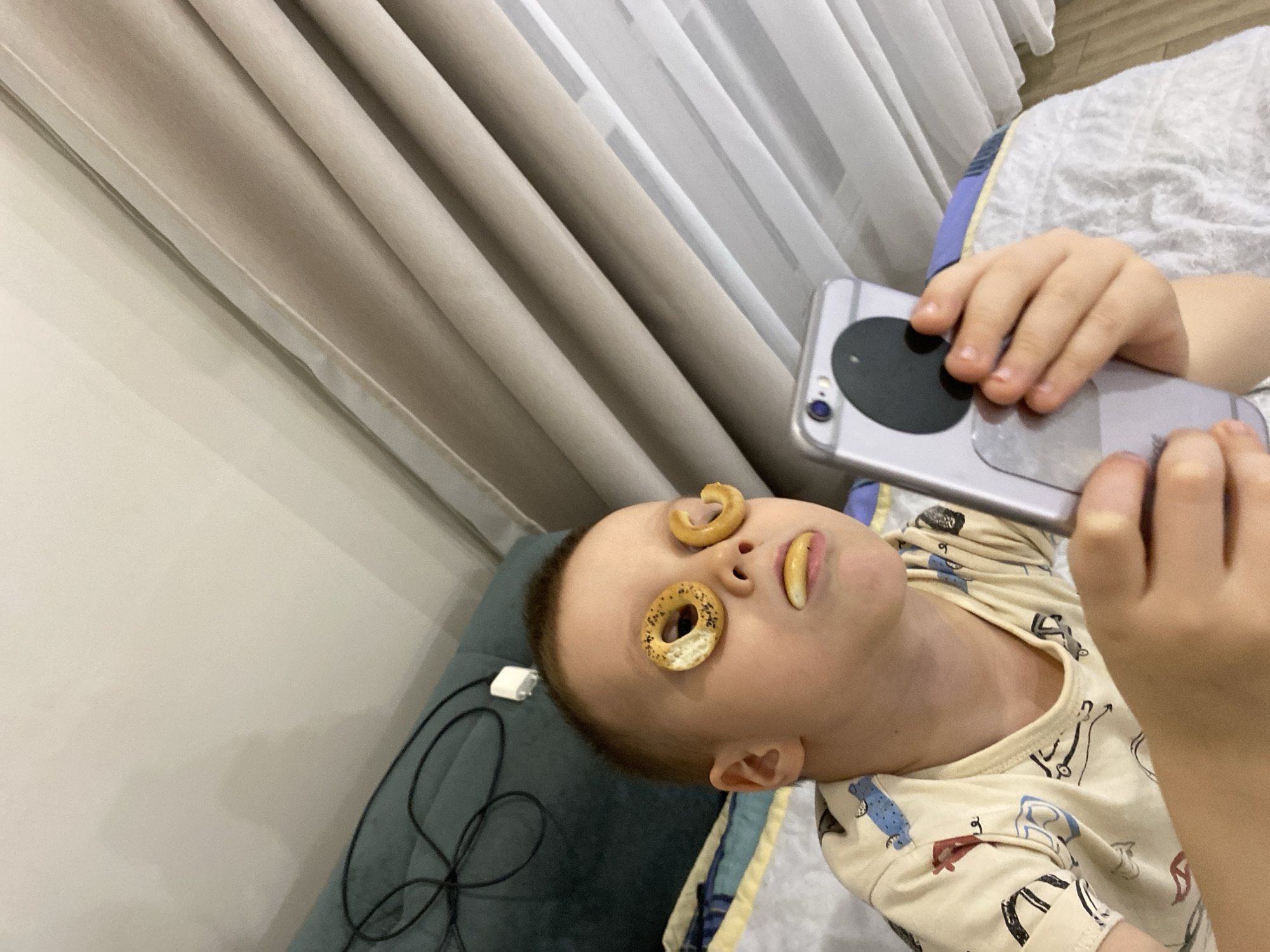 A child lies on a bed with two small bagels on his eyes and a small bread stick in his mouth. He is holding a silver smartphone. He is wearing a beige shirt with various patterns. The bed has a white patterned sheet and a blue and yellow patterned comforter. Curtains are visible in the background. The photo was likely taken in Chaikovsky, Russia.