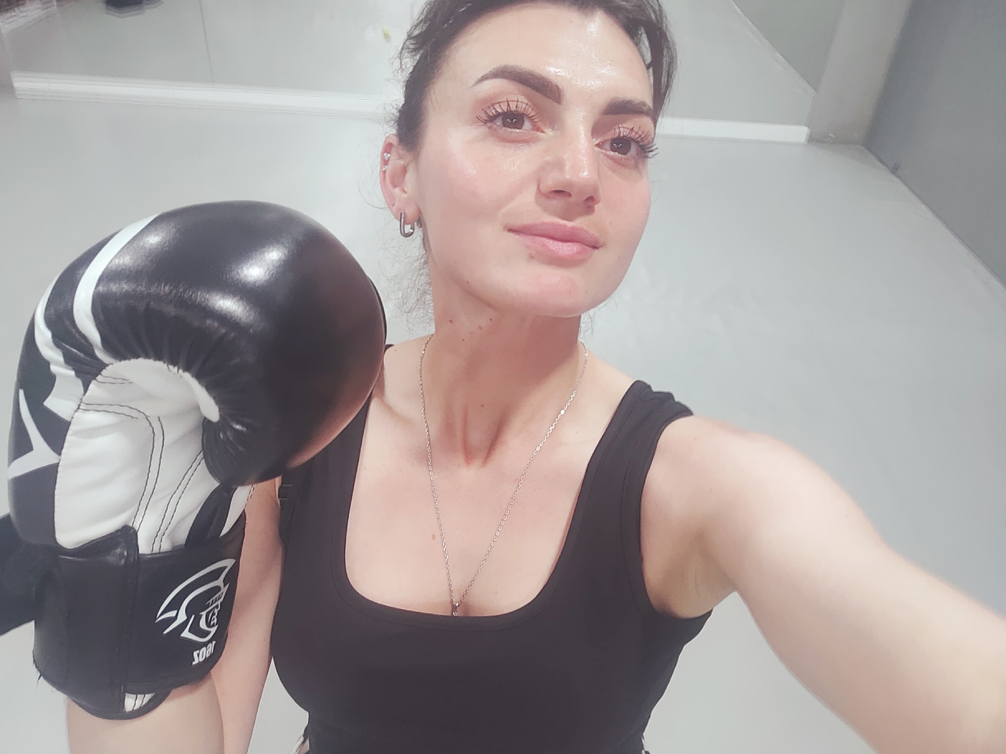 A young woman wearing a black tank top and a silver necklace stands in a gym, holding a black and white boxing glove. She has dark hair pulled back and is wearing hoop earrings. The background is a plain, light-colored wall with some shadows. The image appears to be a selfie, as it is taken from a close-up perspective. The woman is looking directly at the camera with a slight smile.