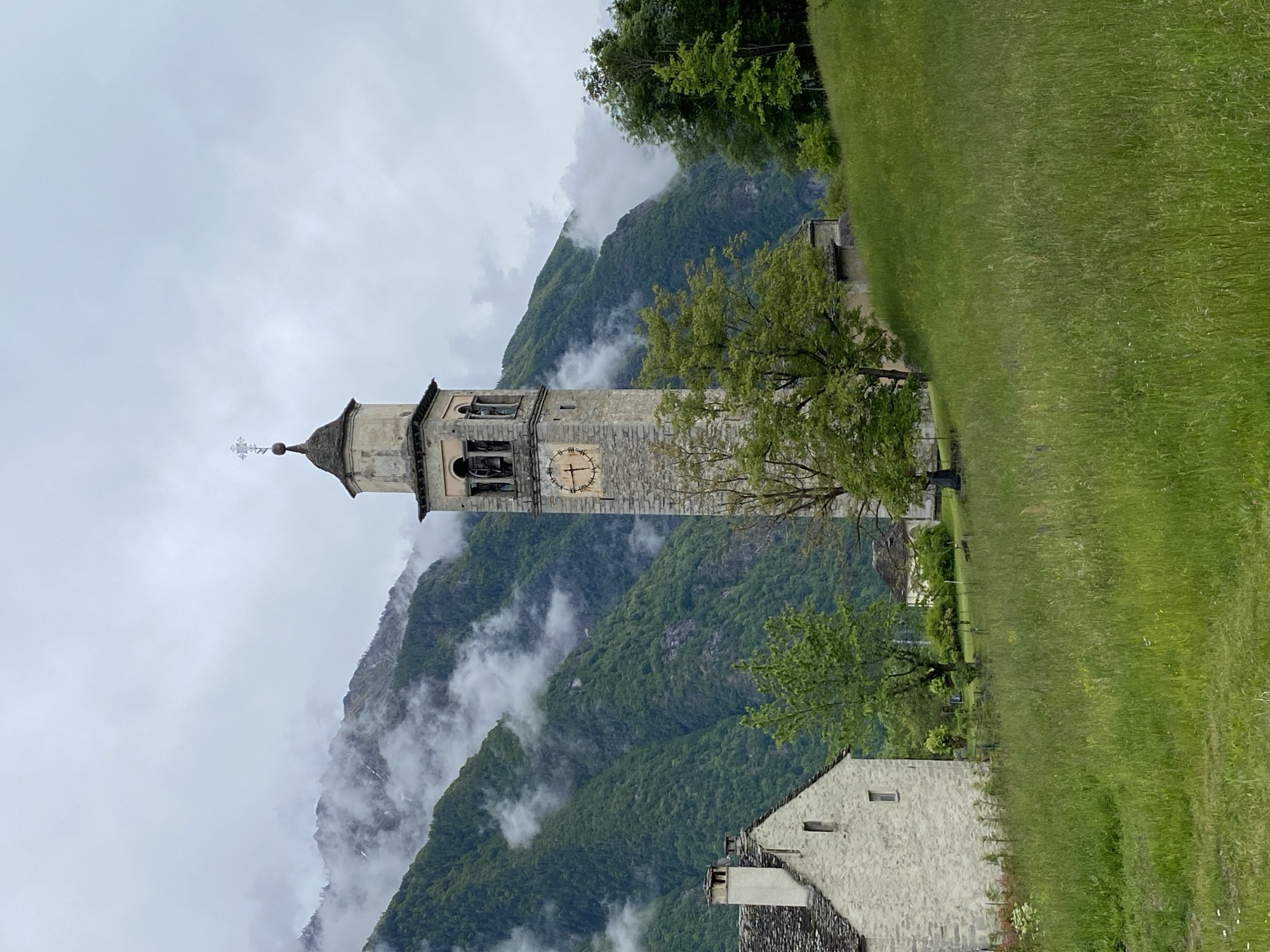 The image shows a stone church tower in Crodo, Italy. The tower is tall and slender, with a clock face visible on its side. The tower is surrounded by a mountainous landscape with fog. In the foreground, there is a grassy field. A stone building with a slate roof is located near the tower.