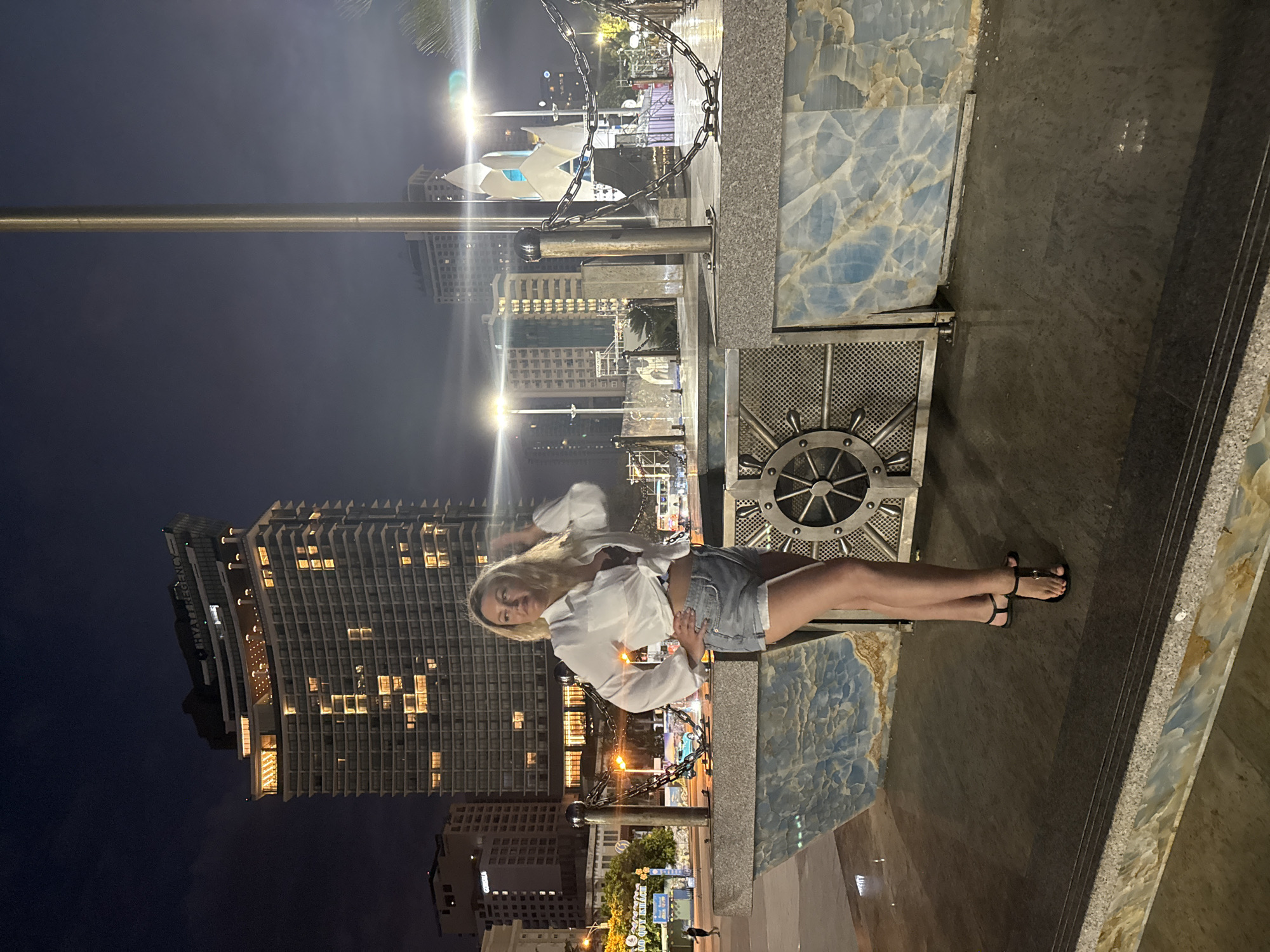 At night, a fair-skinned woman with blonde hair is posing in Nha Trang Ward, Vietnam. She is wearing a white blouse and denim shorts with black sandal footwear. She is leaning against an architectural element that features a wheel design and decorative blue marble-like panels. The background includes city buildings illuminated with lights. In the distance, there are structures and light poles with decorative lighting. A chain-link barrier is partially visible.