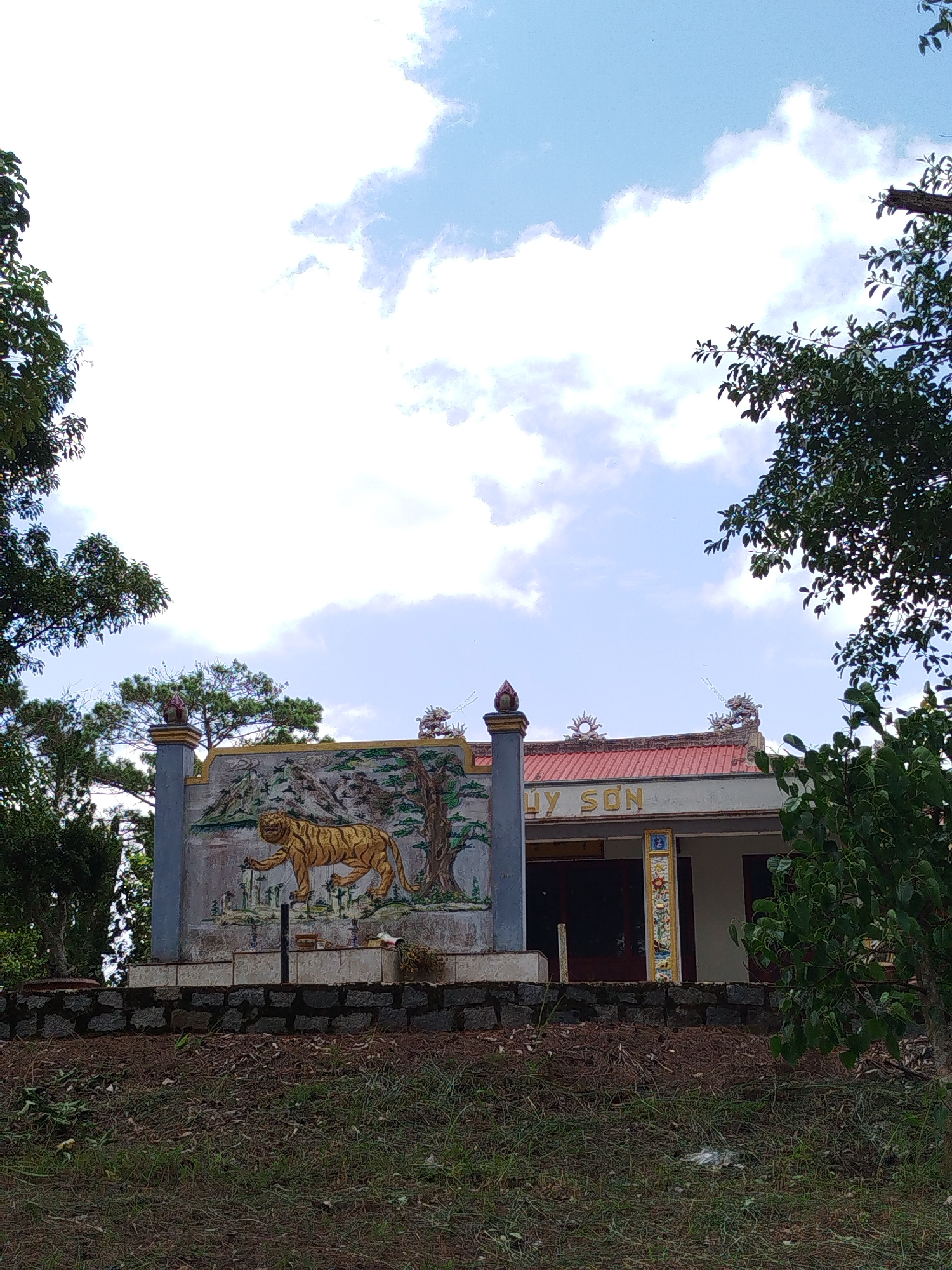 The image shows a building with a red roof and a white facade, possibly a temple or shrine, located in Tuý Sơn, Vietnam. Above the entrance, the words "HUÝ SON" are visible in light-colored lettering. The building is adorned with decorative elements, including a prominent mural on the left depicting a golden tiger walking through a landscape with trees and mountains. The mural is mounted on a grey pillar and a tiled base, which is surrounded by a dark stone wall. Several trees with lush green foliage frame the scene, with branches extending into the upper portion of the image. The sky above is a mix of bright white clouds and patches of blue. The foreground consists of a grassy slope with dry leaves and scattered debris.
