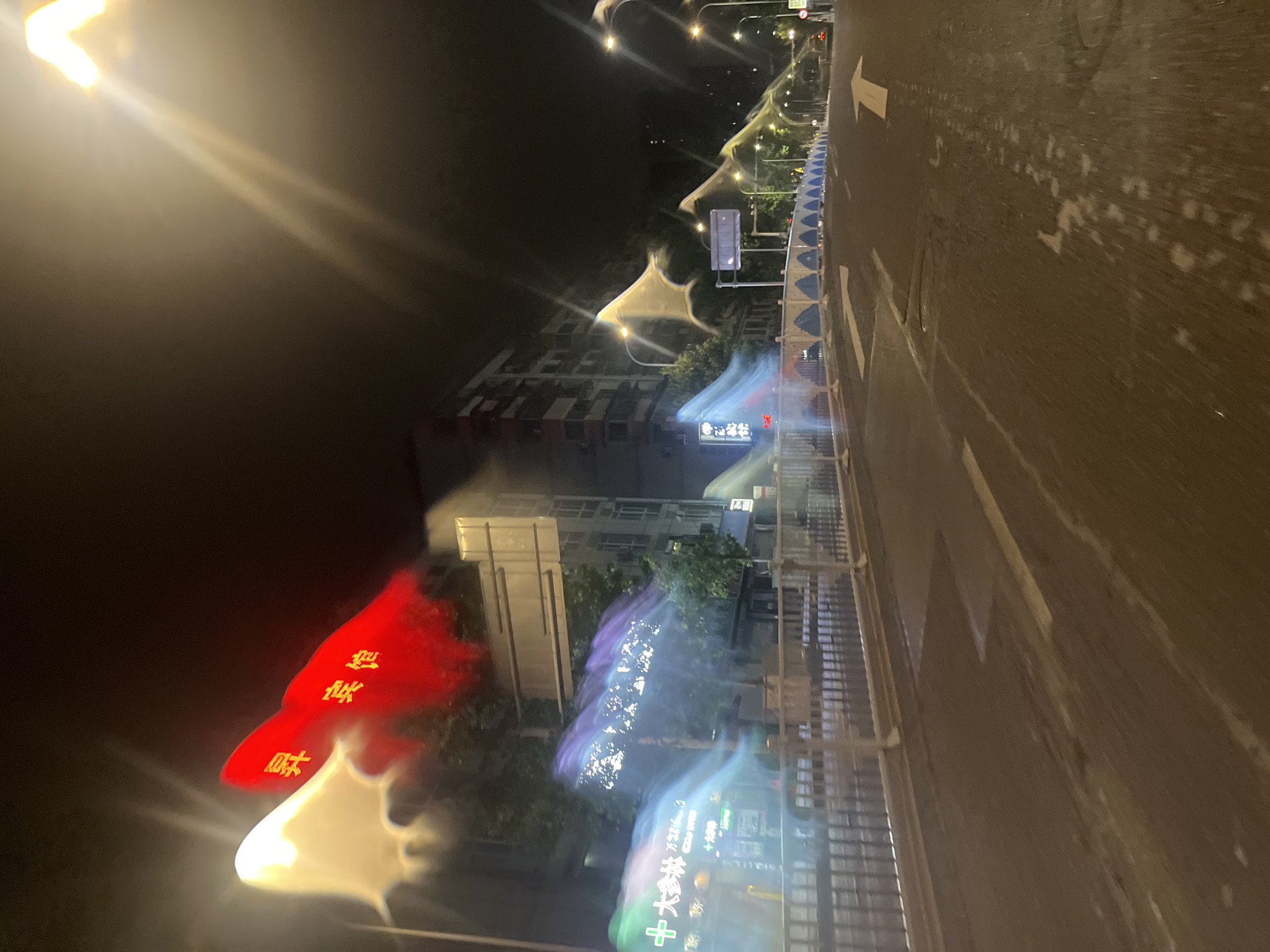 This is a nighttime photograph of a street in Yuzhong District, China, taken from a moving vehicle. The image is characterized by motion blur and light trails, suggesting a long exposure or rapid movement. In the foreground, a dark asphalt road is visible with white directional arrows painted on it, indicating lanes for traffic. The road surface appears wet or reflective in places.
To the left of the road, a white metal railing separates the roadway from a sidewalk or pedestrian area. Behind the railing, several buildings are visible. The most prominent building in the middle ground is a multi-story structure with visible windows. To the left of this building, a brightly lit sign with green and white Chinese characters, including a cross symbol, is partially obscured by motion blur.
Above the street, several objects are illuminated by artificial light, creating abstract shapes and streaks of color. Two of these objects, in the upper left quadrant, appear to be red and white banners or flags with Chinese characters, glowing intensely and blurred. The sky is dark, and streetlights with visible lamps are intermittently spaced along the right side of the road, casting streaks of light that blend with the overall motion blur. The overall impression is one of a dynamic urban environment at night, with vibrant lights and movement.