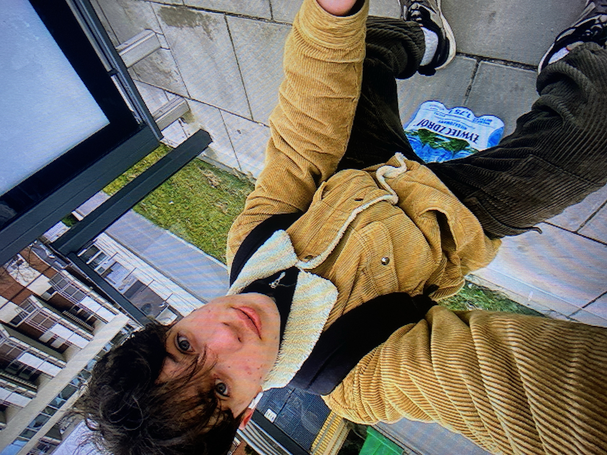 A young person with dark, messy hair and blue eyes is taking a selfie outdoors. They are wearing a yellow corduroy jacket with a cream-colored sherpa collar, a black turtleneck, and dark pants. A black backpack is visible. In front of them, on the ground, is a plastic six-pack of "ŻYWIEC ZDRÓJ" bottled water. The background shows a modern building with balconies and a grassy area, suggesting an urban setting. The ground is paved with large square tiles. The scene appears to be in Poznan, Poland, given the Polish text on the water bottle.