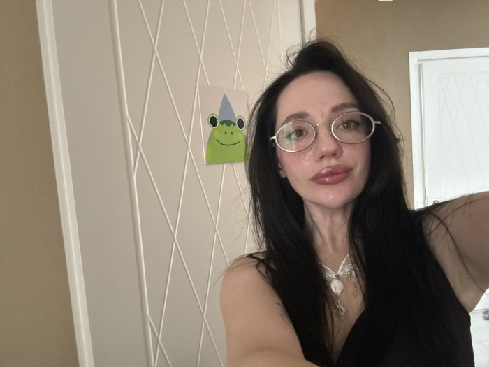 The image presents a close-up indoor self-portrait of an adult female. She has dark, long hair, partially styled in a bun on top of her head, with strands falling over her shoulders. She wears light-rimmed, oval eyeglasses and exhibits visible freckles across her nose and cheeks. Her lips are full and appear to be covered in a light-colored gloss. The subject is wearing a black sleeveless garment and a silver-toned necklace adorned with a central circular pendant and several smaller charms, including star and sun motifs. She holds the camera with her left hand, which is visible in the lower left portion of the frame, and gazes directly at the lens. The background features a white wall with a decorative pattern of intersecting diagonal raised lines. A section of a plain beige wall is visible to the left. A small, rectangular artwork depicting a green frog wearing a blue and white party hat is affixed to the white wall. A white door, featuring a similar geometric pattern as the wall, is partially visible on the far right. The specific location within the city of Batumi, Georgia, cannot be determined from the visual information provided in the image.