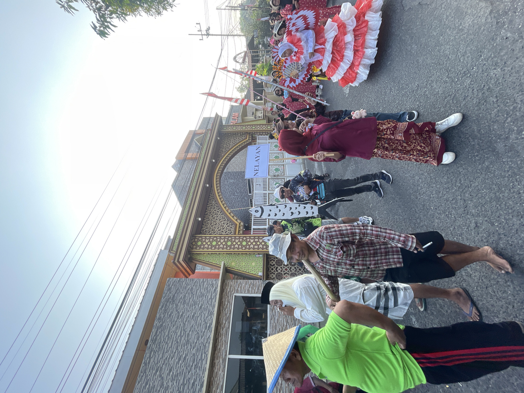 The image captures a street parade in the city of Candi, Indonesia. The sky is clear and bright, indicating daytime. A group of people are walking down a paved street, some in traditional attire and others in modern clothing. In the foreground, a man wearing a lime green shirt and black pants with red stripes is visible from the waist down. To his right, another man in a plaid shirt and black shorts holds a wooden pole. Further back, a person dressed as a fish, holding a sign that reads "NELAYAN", is part of the procession. The background features a building with an ornate, arched entrance, adorned with intricate patterns and green accents. Flags and banners are visible throughout the parade, adding to the festive atmosphere. Several individuals are wearing elaborate costumes, including one in a large red and white ruffled dress. The overall scene depicts a community event or celebration.