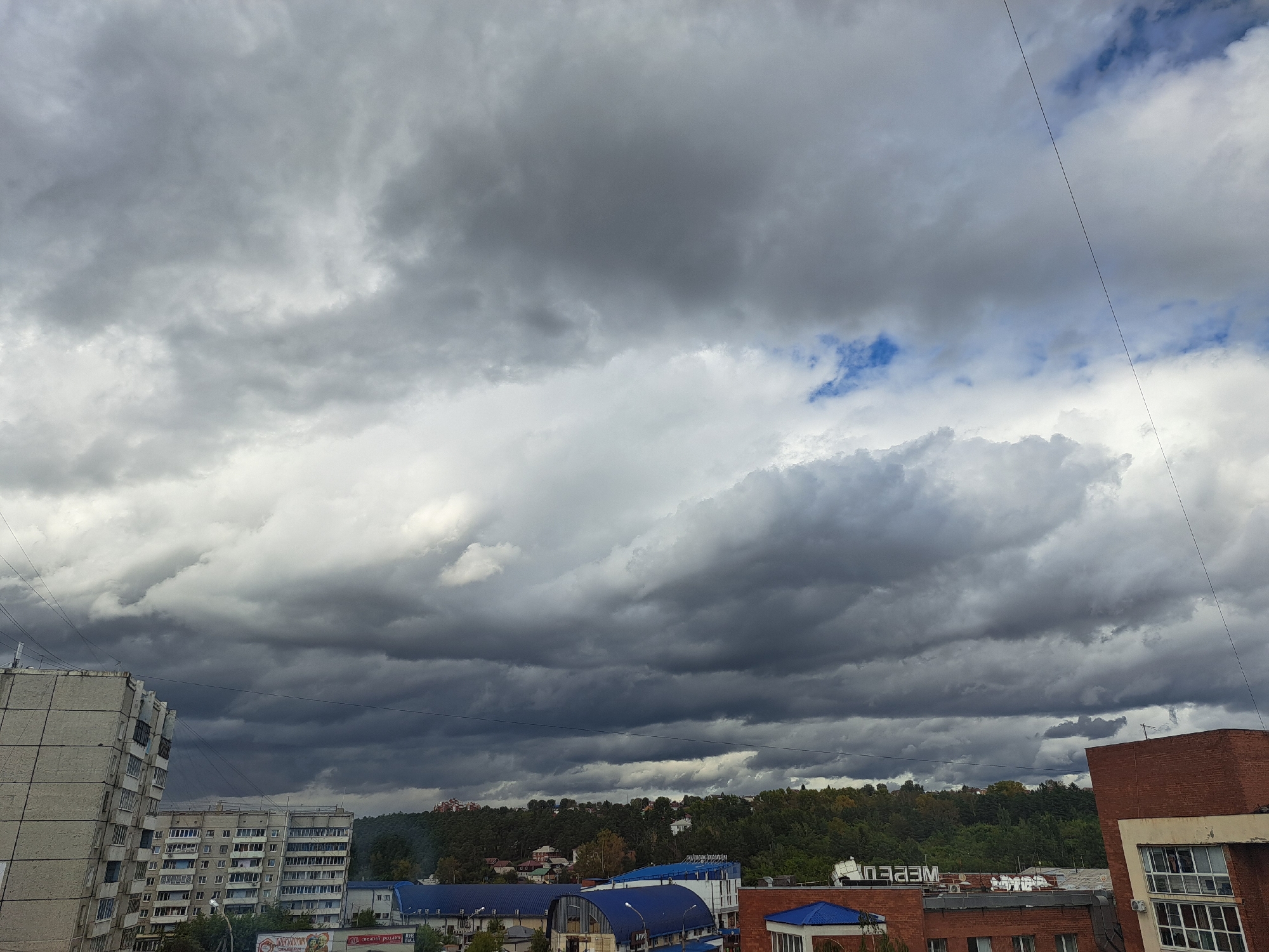 The image captures an overcast sky filled with dark, heavy clouds, indicative of impending rain or a storm, over a section of the city of Irkutsk, Russia. The foreground features several multi-story residential buildings constructed from concrete, with numerous balconies and windows. Adjacent to these residential structures, there are commercial or industrial buildings with blue and white roofs, and one prominent brick building. Beyond the immediate urban landscape, a densely wooded hill serves as a backdrop, with some scattered houses visible amidst the foliage. Power lines are visible crossing the sky. No people are discernible in the image, and no specific actions are being taken. The environment appears to be a typical urban scene on a cloudy day.