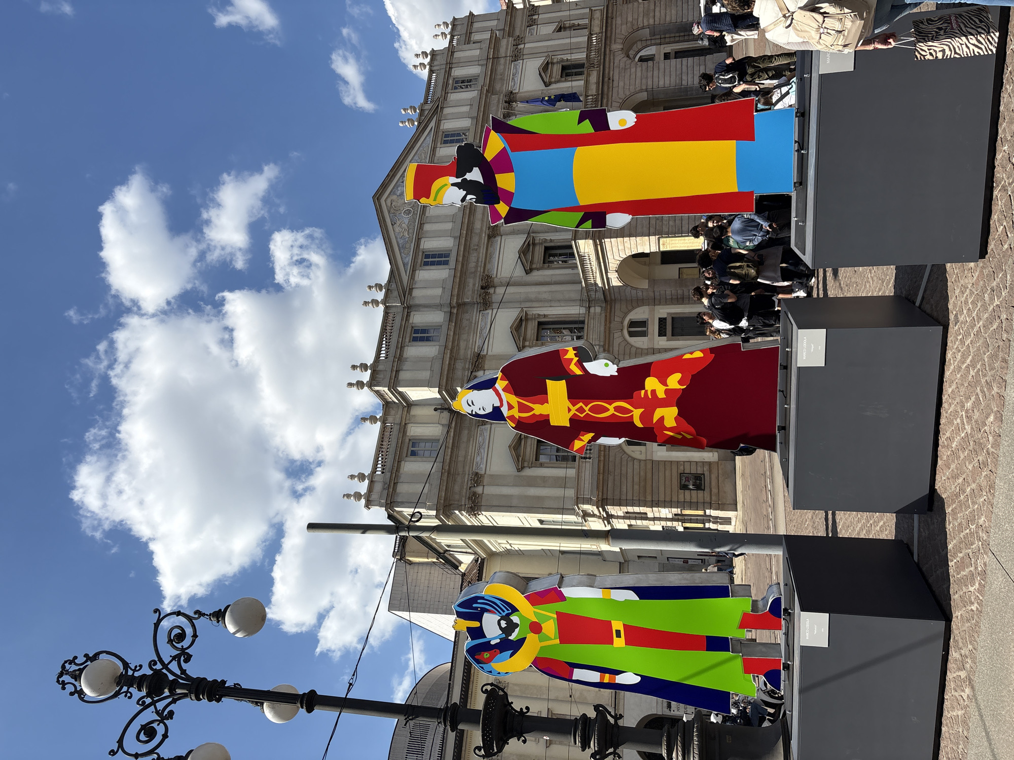 The image shows an outdoor art installation in what appears to be a public square in Milan, Italy. There are three large, brightly colored figures resembling stylized historical or medieval characters. The figures are displayed on dark gray plinths. Behind the art installation is a multi-story building with a classical facade. The sky above is blue with scattered white clouds. A wrought-iron lamppost is visible on the left side of the frame. In the background, there are people gathered near the installation. The image is taken from a low angle, emphasizing the height of the figures and the building behind them. The light suggests it is daytime.