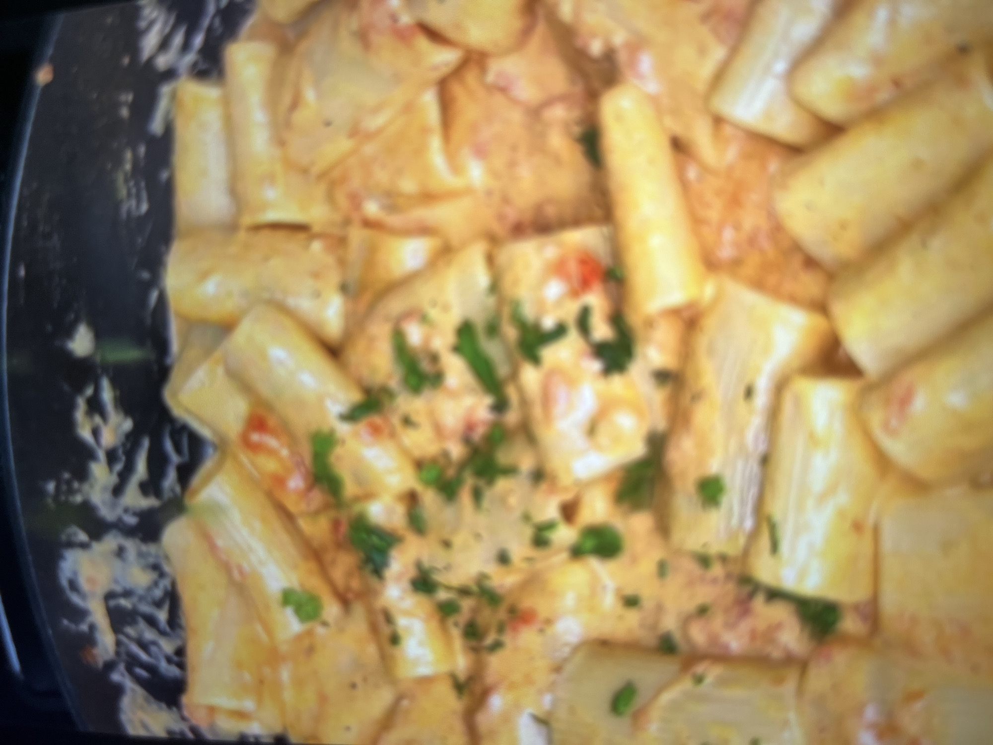 The image shows a close-up, overhead view of a dish of rigatoni pasta in a creamy, orange-colored sauce, garnished with chopped parsley. The pasta pieces are large and tubular, and they are mixed with small red pieces, likely tomatoes or peppers, and flecks of green herbs. The sauce appears rich and smooth, coating the pasta generously. The dish is served in a dark, shallow pan or pot. The lighting is somewhat dim, and the image has a slightly blurry, low-resolution quality. There is no visible indication of the location in the city of Abuja, Nigeria.