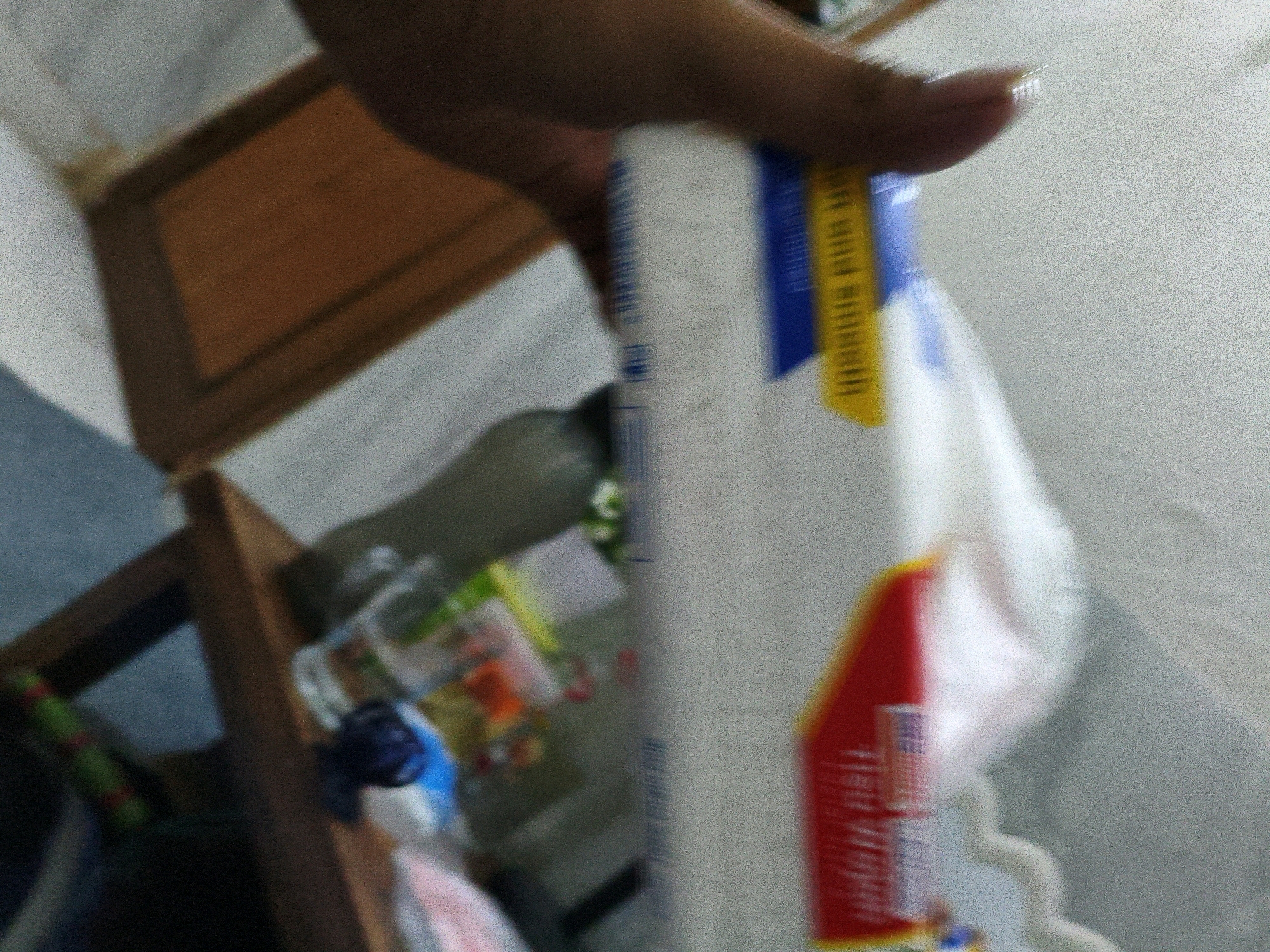 The image shows a blurry indoor scene. In the foreground, a person's hand is holding a white object with blue and yellow labels, possibly a cleaning product or dispenser. The labels have text that is unreadable due to the blur. In the background, there are wooden shelves and some bottles, which are also blurry. The lighting suggests an indoor environment. There is no identifiable information within the image to associate it with Karangtalun, Indonesia.