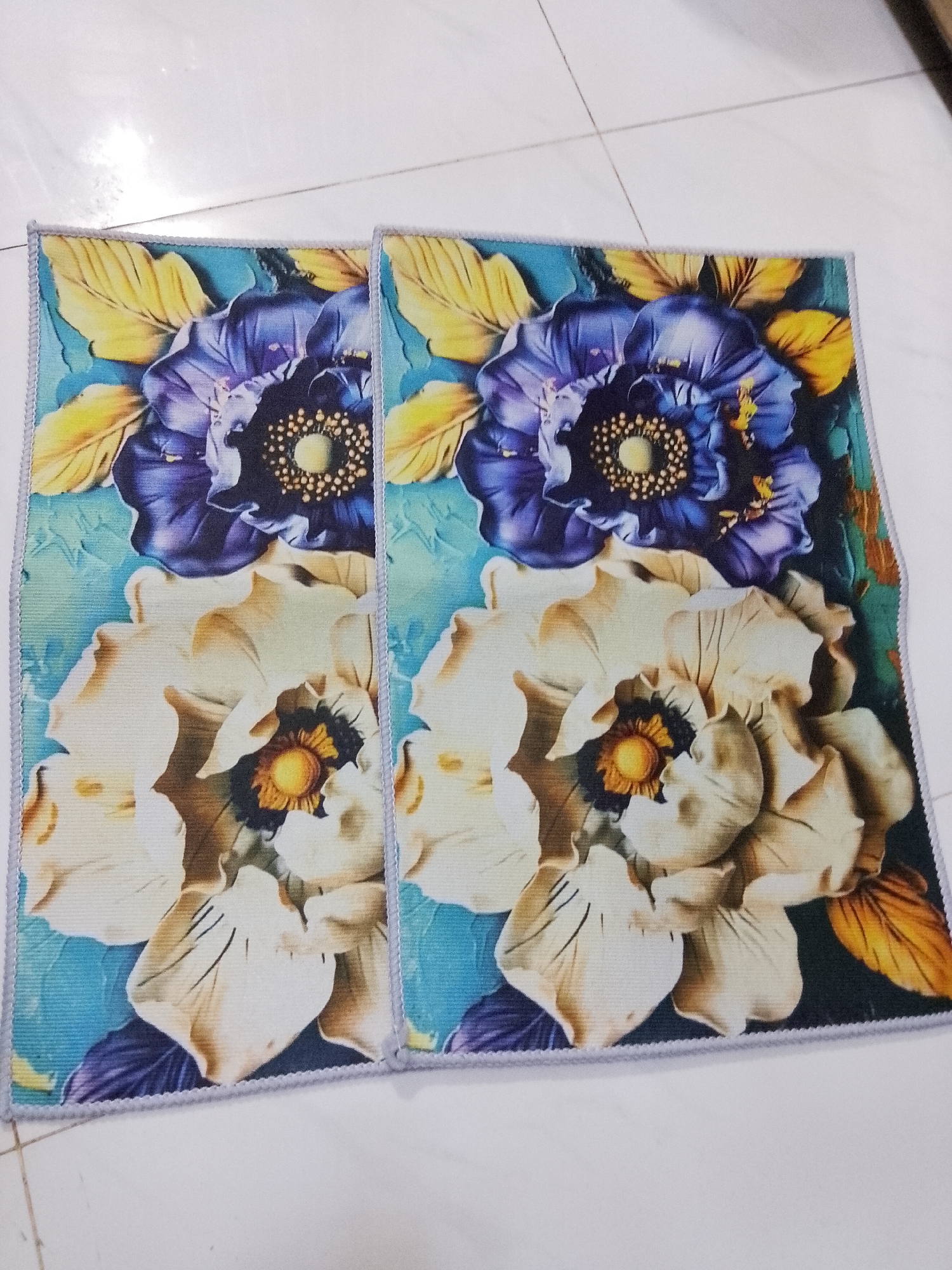 The image captures two identical square-shaped mats or small rugs, positioned side-by-side on a light-colored, possibly white or off-white, tiled floor. The setting appears to be indoors during the daytime, indicated by the bright ambient lighting.
Each mat features a vibrant, somewhat painterly floral design that gives the impression of a 3D or embossed texture. The primary subjects of the design are two large flowers: a prominent deep purple or indigo flower situated towards the top, and a large cream or off-white flower positioned below it. Both flowers exhibit a distinct central area with yellow or orange hues, surrounded by numerous small dots or stamen-like patterns. The background of the floral motif is a blend of blue and teal, interspersed with several yellow-orange leaves that complement the main blossoms.
The edges of both mats are finished with a light grey, braided-look border. The tiled floor beneath the mats is clean and reflects some light, typical of indoor flooring. There is no activity taking place; the image is a static shot of the objects. No people or animals are present, and consequently, no emotions are conveyed. There is no visible text within the image. The photograph is taken from a slightly elevated angle, looking down onto the mats.