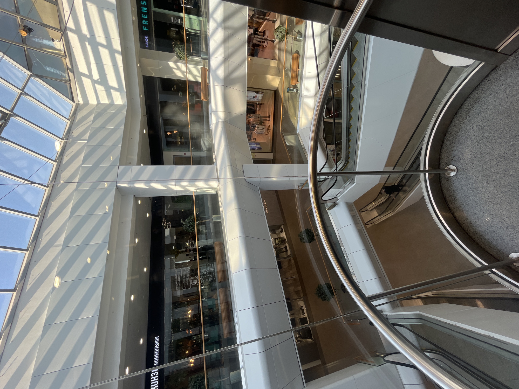 The image depicts the interior of a shopping mall, likely located in Izhevsk, Russia based on the presence of Cyrillic script in the signage. The view is from an elevated position, looking down into a multi-level atrium. The architecture is modern, with white structural columns and glass-fronted retail spaces stacked vertically. Sunlight streams through a skylight, casting shadows across the walls. Escalators connect the various levels. There are a few people visible, mostly as small figures in the distance. The foreground features a metal railing, indicating the vantage point is from a walkway or balcony. The general atmosphere is bright and spacious.