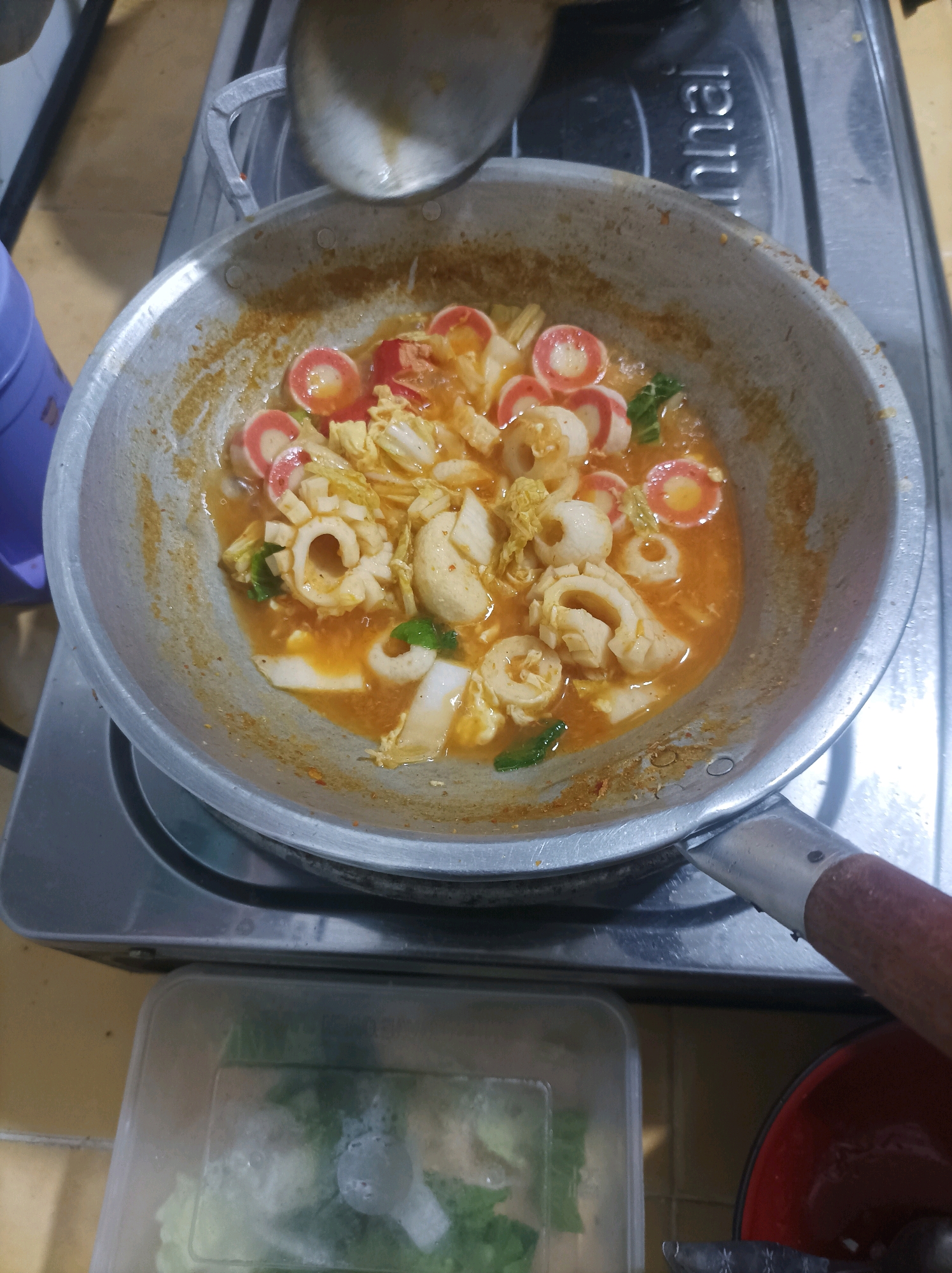 An overhead, medium shot captures a wok filled with a saucy dish containing sliced squid rings, what appear to be fish balls, and Napa cabbage, all simmering in an orange broth. The wok is made of aluminum and shows signs of use, with some browning and residue on the interior surface. A wooden-handled ladle is partially visible at the top of the frame, as if in the process of stirring the dish. The wok rests on a grey gas stove, which has the word "Rinnai" imprinted on it. Below the stove, a clear plastic container holds chopped green vegetables. To the right of the container, a red bowl is partially visible. The setting appears to be a kitchen in Canga'an, Indonesia, with the overall scene conveying a sense of home-cooked meal preparation.