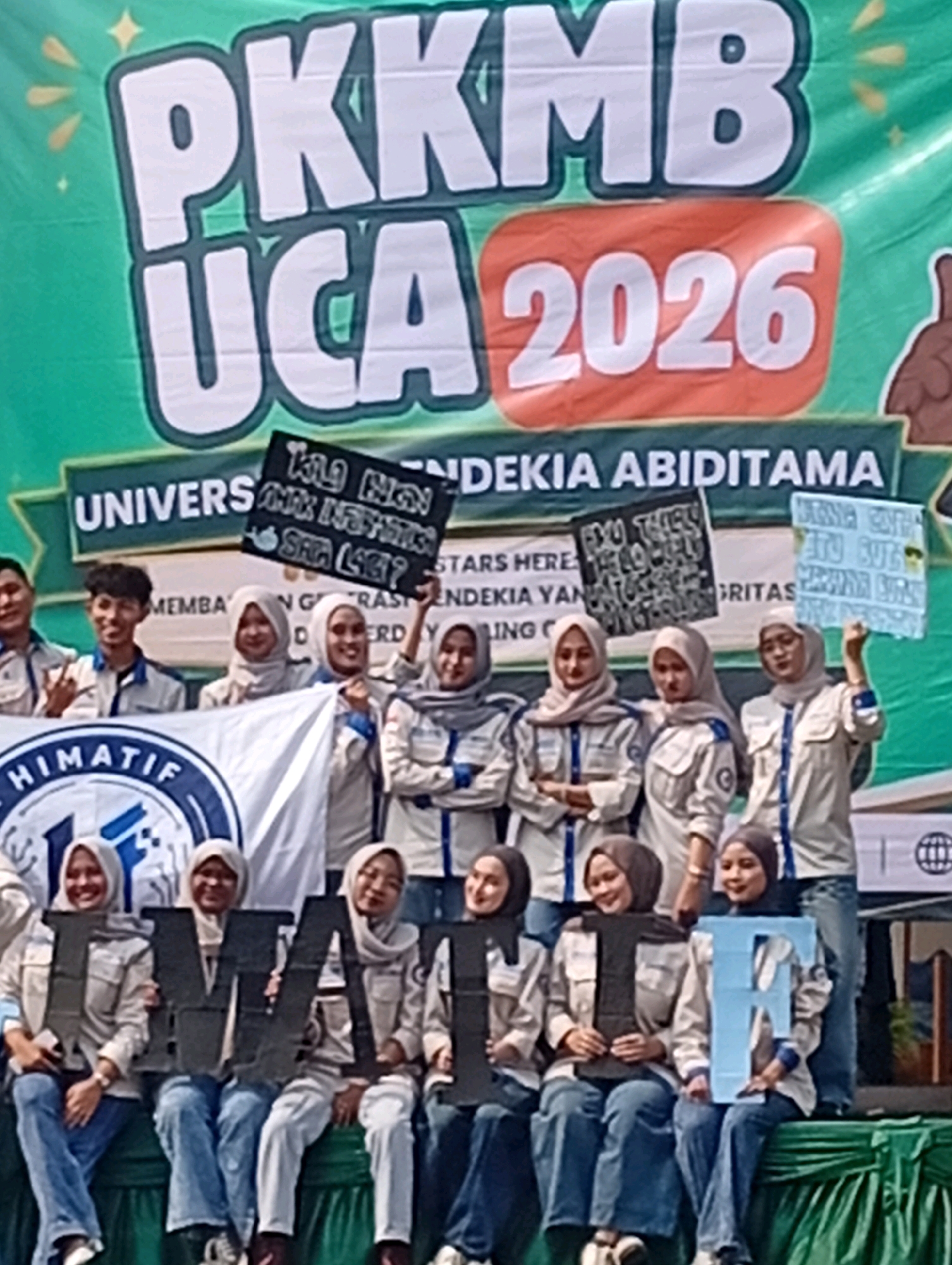 A group of young adults, predominantly women wearing headscarves and matching uniforms, are posing for a photograph in front of a large green banner. The banner has bold white lettering that reads "PKKMB UCA 2026." Below this, it says "UNIVERSITAS TENDEKIA ABIDITAMA" in smaller white text. Several individuals are holding up black and light blue signs with writing on them. Some of the people are also holding large, dark gray, and light blue letters that spell out "IMAFT." The individuals are smiling and appear to be participating in an event or orientation. The background suggests an outdoor setting, possibly a campus or public space, with some foliage and a building structure visible. The clothing and the banner indicate a formal event related to a university. The event is likely taking place in Kelapa Dua, Indonesia, given the university's name.
