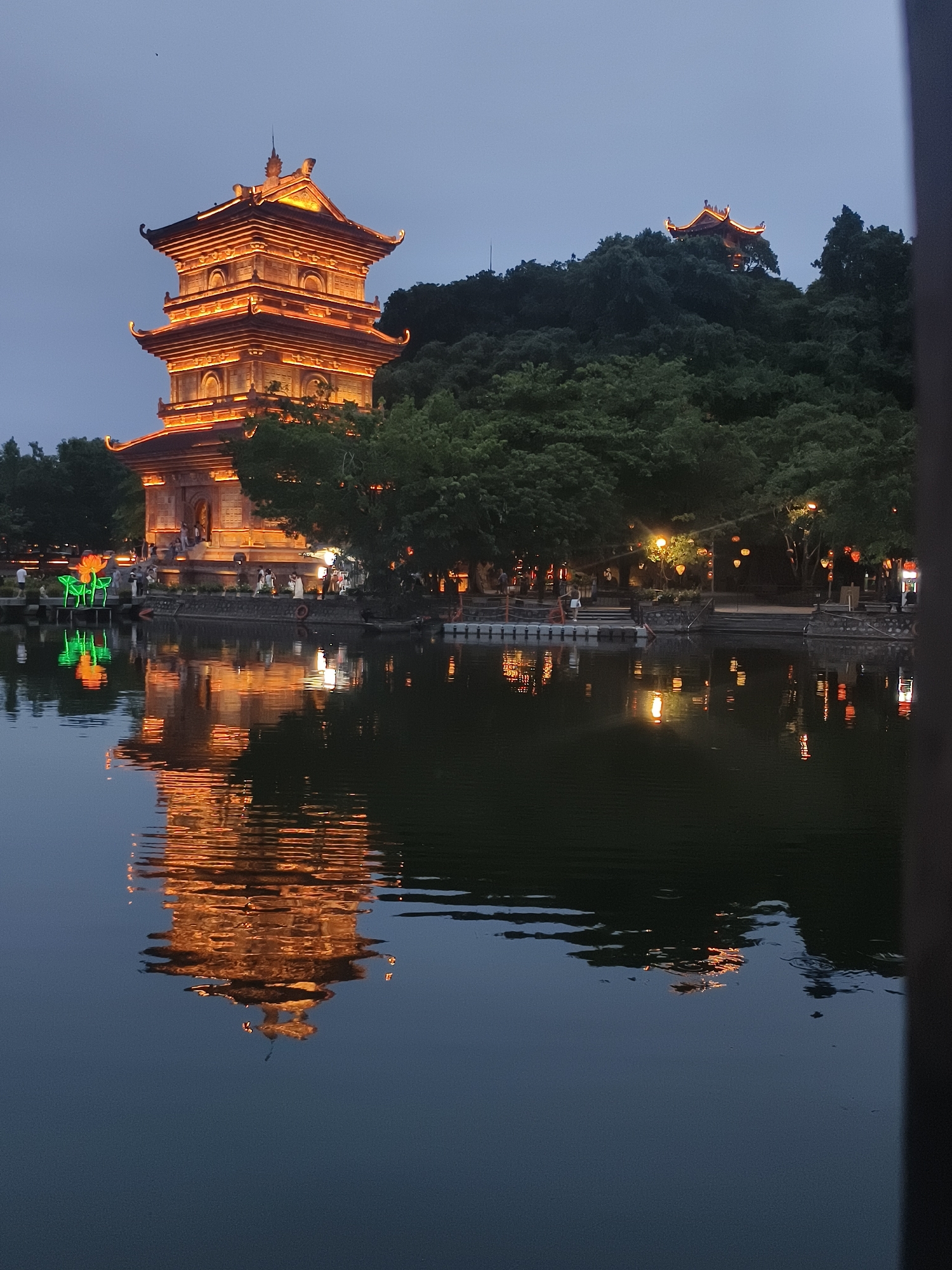 The image depicts a pagota and its reflection in a body of water, likely a lake, in the city of Hoa Lư Ward, Vietnam. The pagota, illuminated with warm orange lights, stands tall and features multiple tiers with upturned eaves, characteristic of traditional East Asian architecture. The surrounding area is characterized by lush green trees. In the foreground, the calm water reflects the pagota and the surrounding lights, creating a symmetrical and serene scene. The sky is a dusky blue, suggesting it is either dawn or dusk. There are a few individuals visible near the base of the pagota and along the waterfront, engaged in leisurely activities. Decorative lanterns are also visible in the background, adding to the ambient lighting and festive atmosphere.