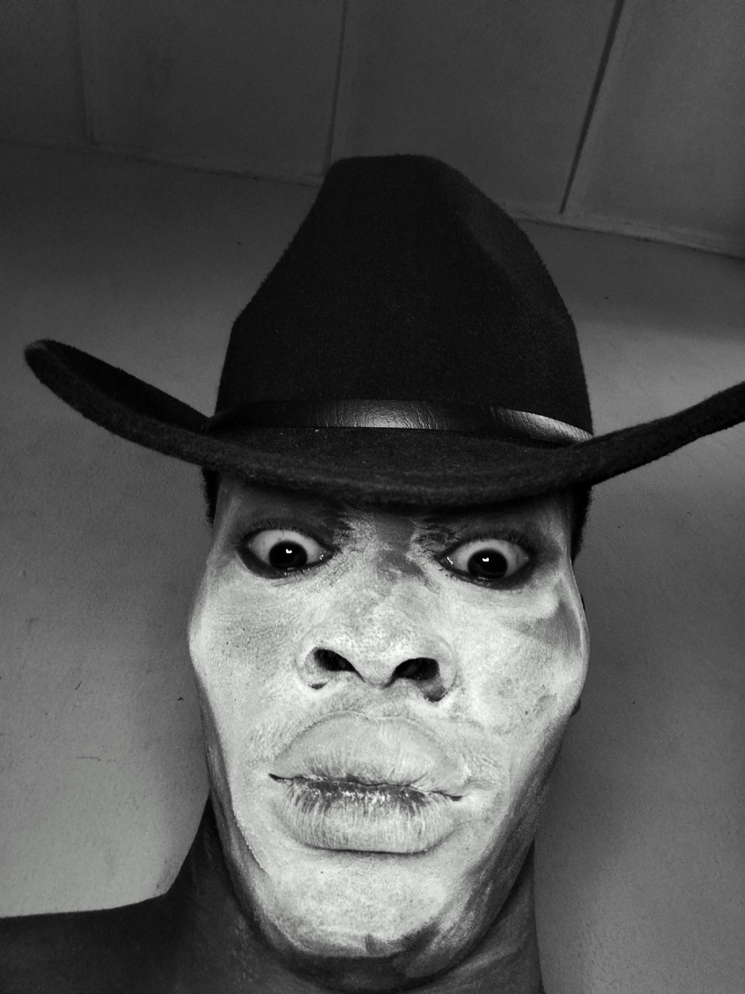 In a close-up, black and white, front-facing selfie, a person with their face painted white and wearing a black cowboy hat is the sole subject. The individual's eyes are wide open, and their mouth is slightly parted. The background consists of a plain, textured wall. The image was taken in Sabon Tasha, Nigeria.