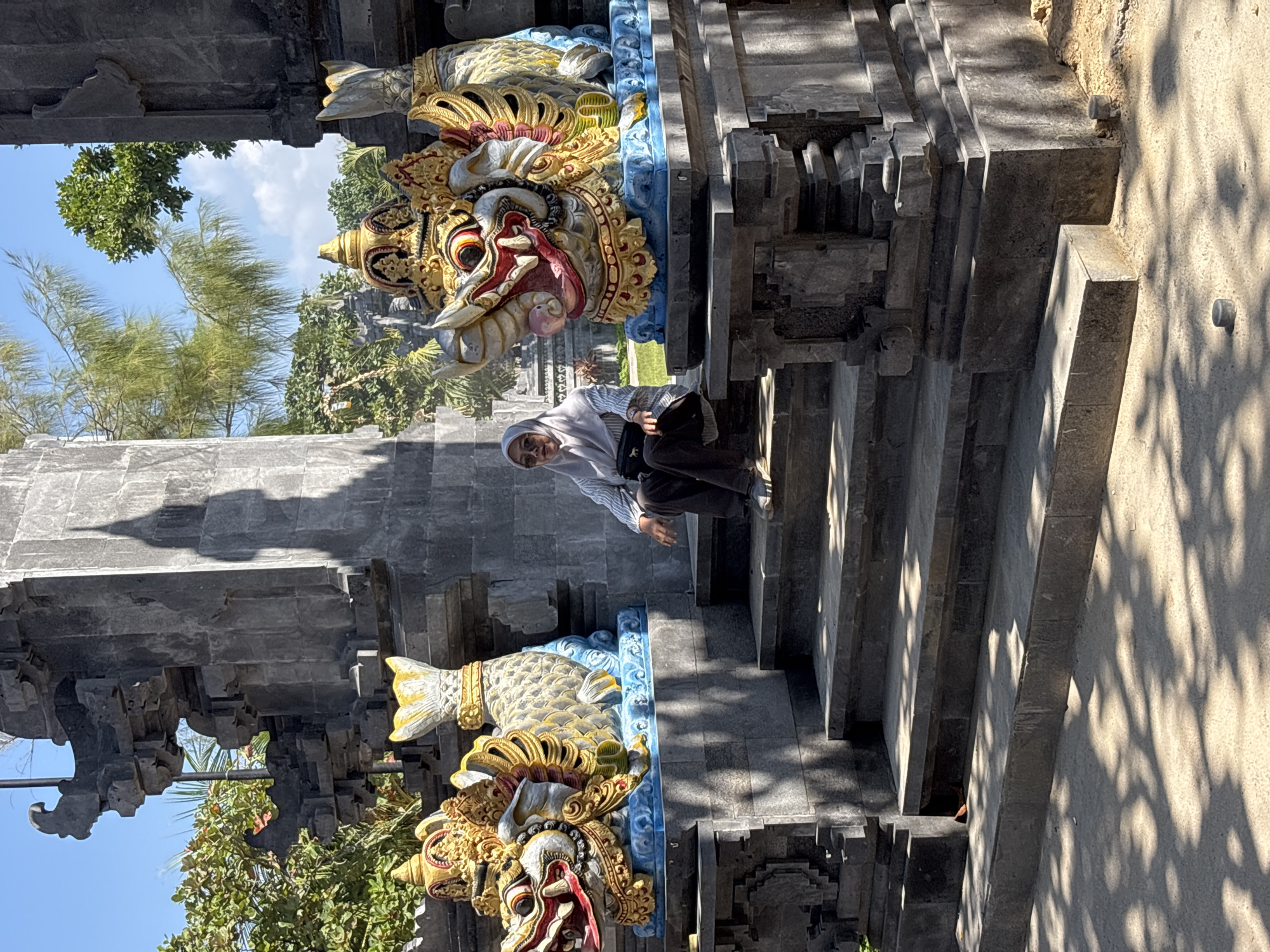 A woman wearing a light grey hijab and a striped long-sleeved shirt sits on the stairs in front of a large stone structure. She is wearing dark pants and grey shoes. To her left and right are colorful, ornate statues of mythical creatures, each with a dragon-like head and a fish-like body. The statues are perched on blue platforms. The stone structure appears to be an entranceway or archway, with intricate carvings and a weathered appearance. The ground in front of the stairs is covered in sand and dappled with sunlight and shadows from trees. The background shows lush green trees and a bright blue sky. The scene suggests a cultural or religious site, likely in Kuta Selatan, Indonesia, given the architectural style and decorative elements.