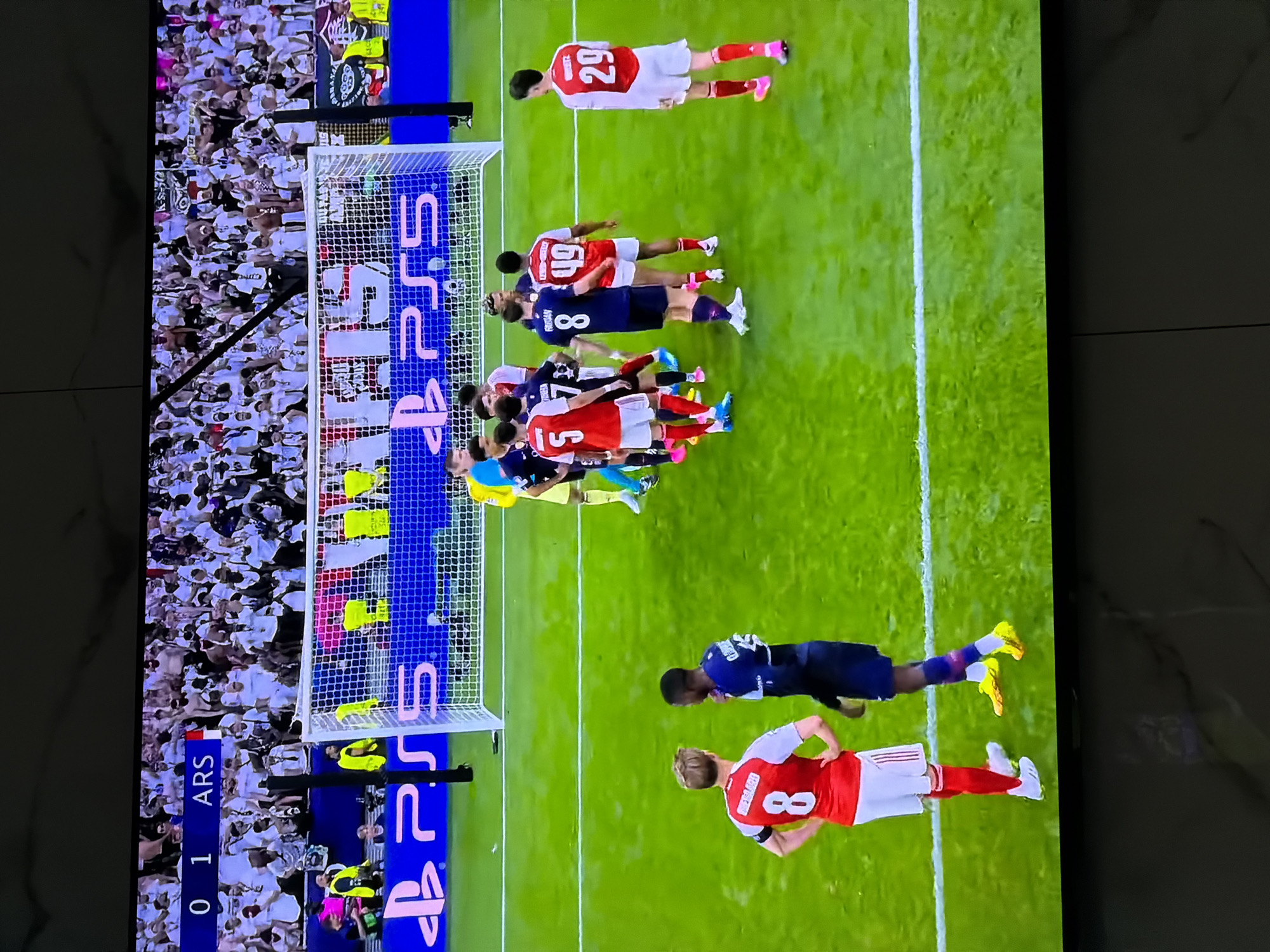 The image shows a televised soccer match. The score is 0-1 in favor of Arsenal (ARS). A free kick is set up with players from both teams positioned near the goal. A group of Arsenal players is forming the defensive wall. Another Arsenal player, wearing number 8, is standing behind them. Two players from the opposing team are nearby. The goal is situated to the left, adorned with "PS5" advertising on the net and behind the goal. A crowd fills the background. The image is captured from a television screen. No information is available about the location in the city of Asenovgrad, Bulgaria.