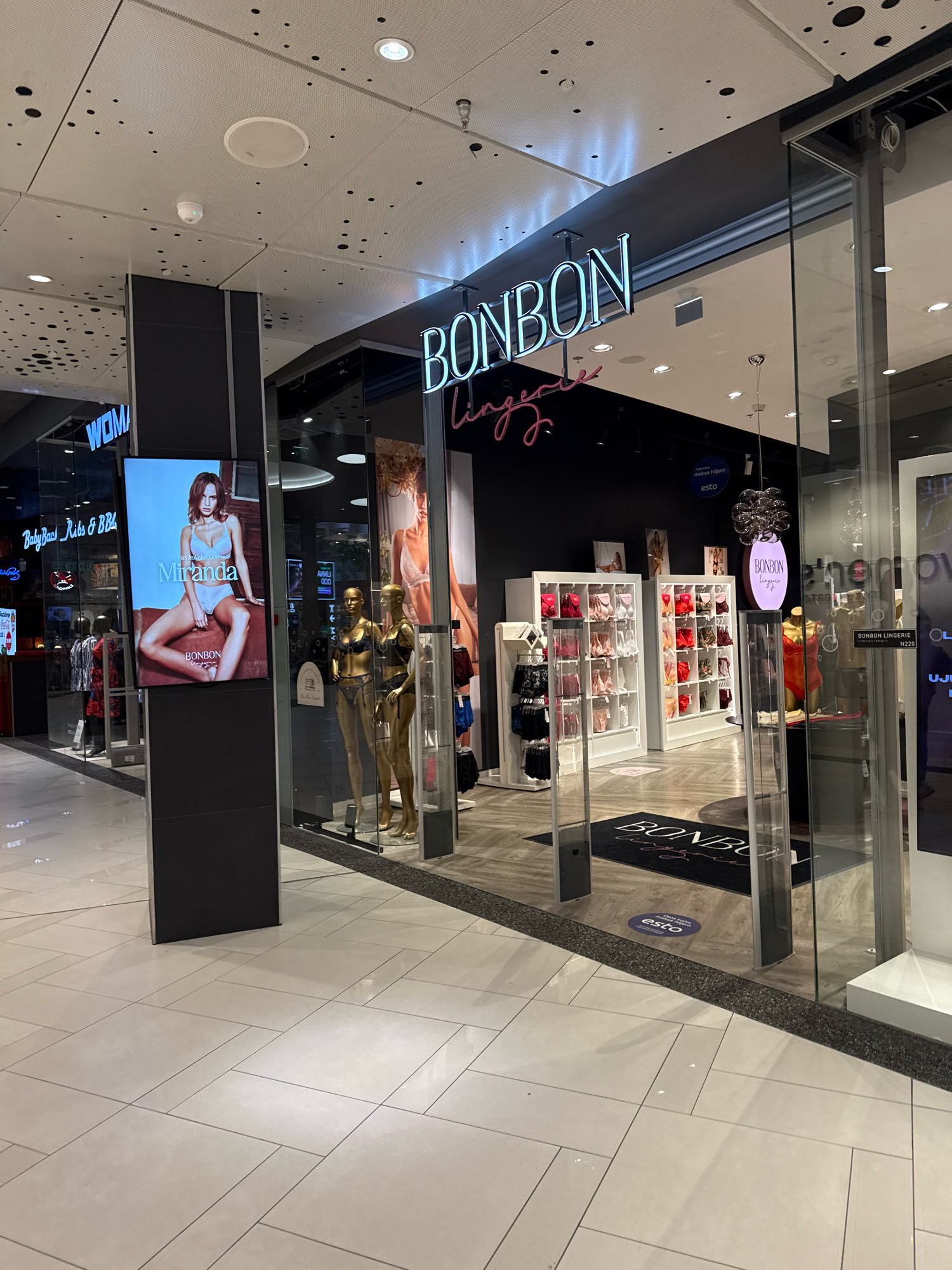 In the city of Tallinn, Estonia, the image shows the entrance to a "BONBON Lingerie" store located within a shopping mall. The store features a large digital display with an advertisement for "Miranda" lingerie, featuring a model in lingerie. Inside the store, mannequins are displayed, some wearing lingerie. Shelving units with various lingerie items are also visible. The flooring appears to be tiled, and the overall environment suggests a retail setting. The lighting is bright, likely from overhead mall lighting and store displays. There are no discernible people actively interacting with the store or each other.