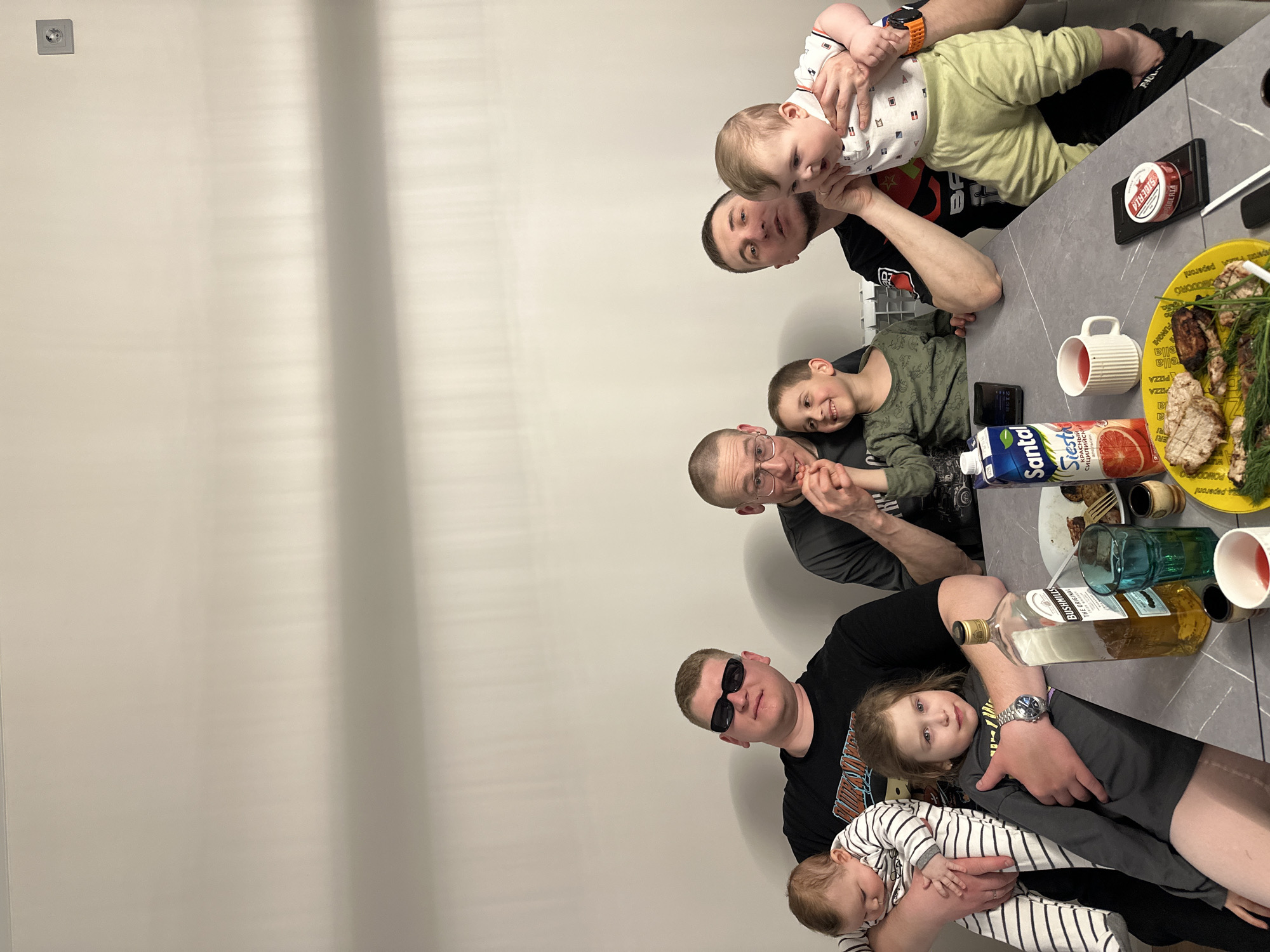 The image depicts six individuals, including four adults and two children, gathered around a table. The setting is likely indoors, suggested by the presence of a wall in the background. From the right and moving counterclockwise, an adult man holds a baby, another adult man is sitting at the table with a child, an older adult man is sitting next to them, and lastly, an adult man is holding a baby. The adults are positioned close to the table. The child is dressed in a black-and-white striped onesie, while another child is wearing green clothing. Food, a bottle of Bushmills whiskey, juice, and a can of Siberia nicotine pouches are on the table. The table and items on it are the visual center of the image, appearing in detail with varied textures and colors. The location is inferred to be in the city of Chaikovsky, Russia, based on the presence of the "Santal Сицилийский красный" (Sicilian red) juice box, the can of "Siberia" nicotine pouches, and other local products which are readily available in Russia.