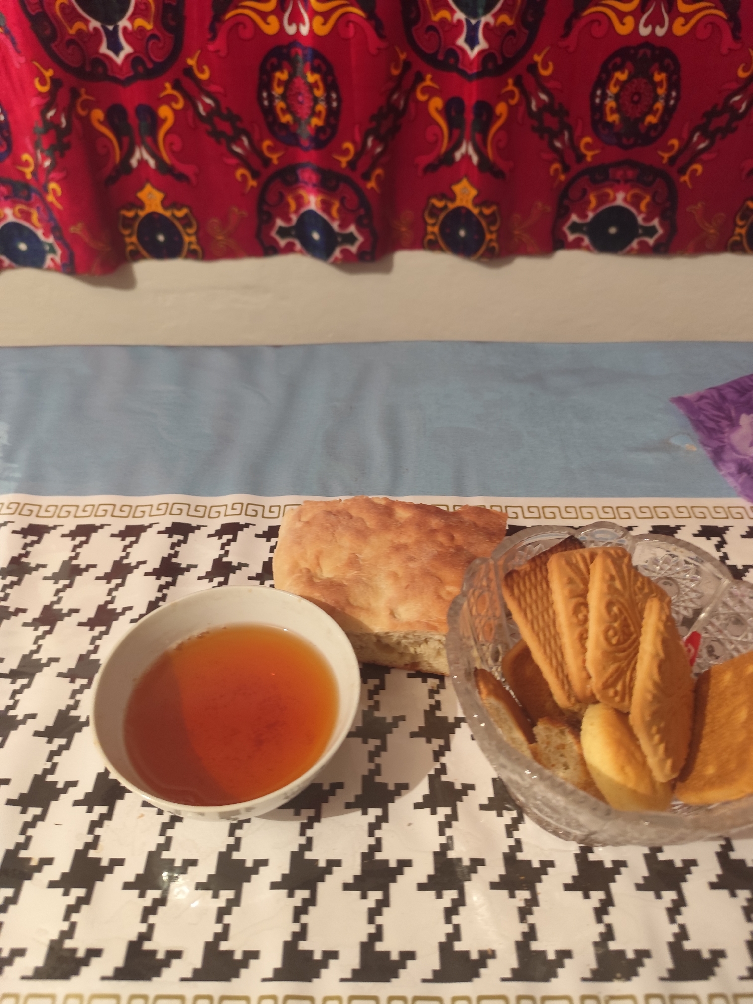 The image shows a close-up of a table setting with a bowl of amber-colored liquid, a piece of flatbread, and a clear glass bowl filled with rectangular biscuits. The table is covered with a patterned cloth featuring black and white houndstooth designs, bordered by a decorative gold pattern. Behind the table, a colorful fabric with a red base and intricate blue, yellow, and white patterns is visible, likely a tablecloth or curtain. The setting appears to be indoors, on a table, possibly in a home. The location is Aqbulaq Rural District, Kazakhstan.