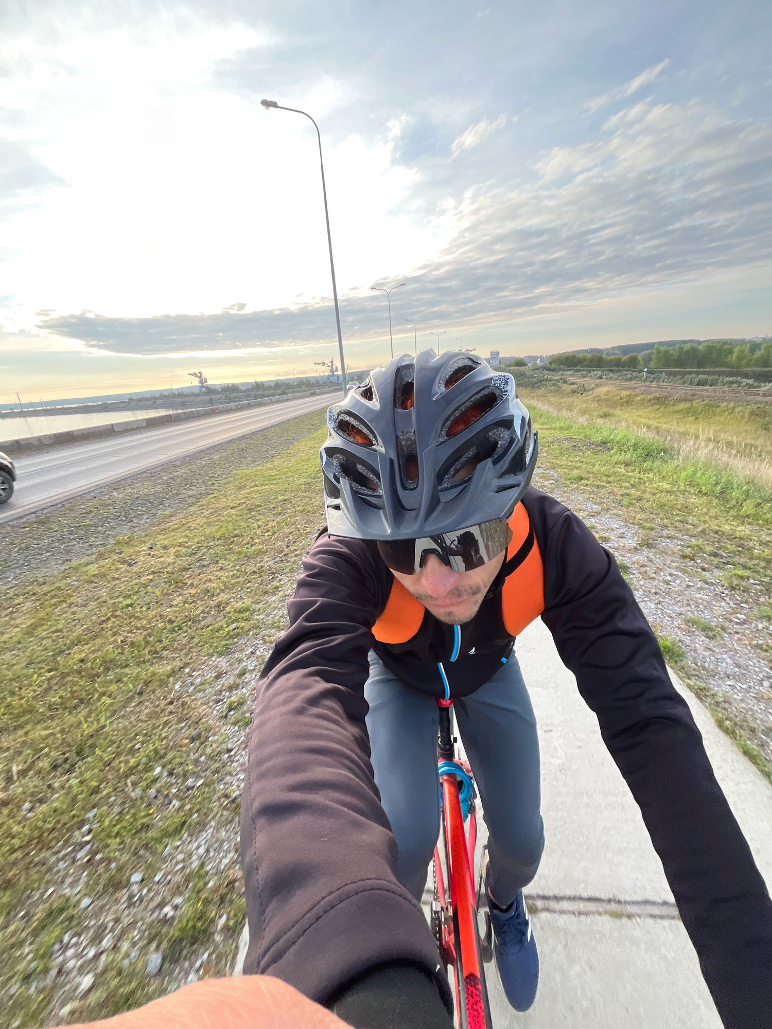 In the city of Chaikovsky, Russia, a first-person perspective shot captures a person riding a bicycle on a paved path adjacent to a road. The cyclist is wearing a dark helmet, sunglasses with reflective lenses, a black long-sleeved jacket over an orange vest, and grey pants. The bicycle has a red frame. The road runs parallel to a body of water on the left, with industrial cranes visible in the distance across the water. To the right of the path is a grassy embankment leading to a railway track and a wooded area with a hazy skyline. The sky is partly cloudy, with the sun creating a bright glare in the upper left quadrant. Streetlights are visible along the roadside. A car is partially visible on the road in the background.