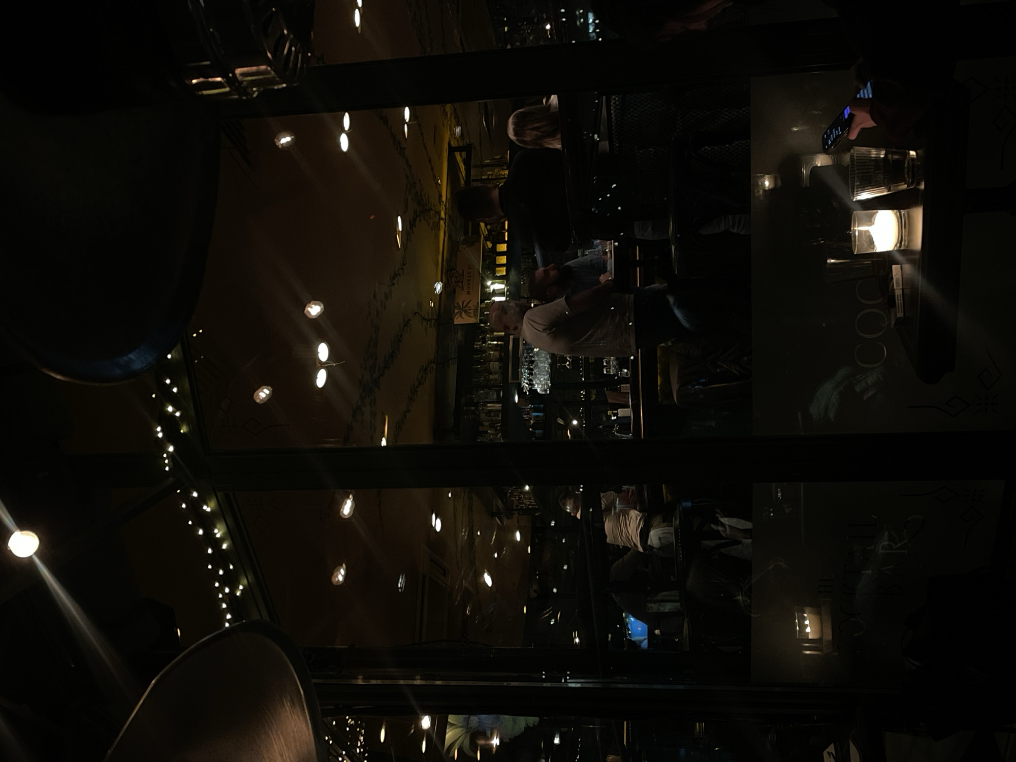 The image shows the interior of a bar or restaurant in Sofia, Bulgaria, taken through a glass partition or window. People are seated at tables and at the bar. In the foreground, parts of chairs are visible. String lights and other light fixtures are visible. A person is holding a phone, possibly taking a photo. Other objects visible on the tables include glasses, candles, and what appears to be a packet of cigarettes. Behind the bar, bottles of alcohol are displayed. Wall decor is also visible.