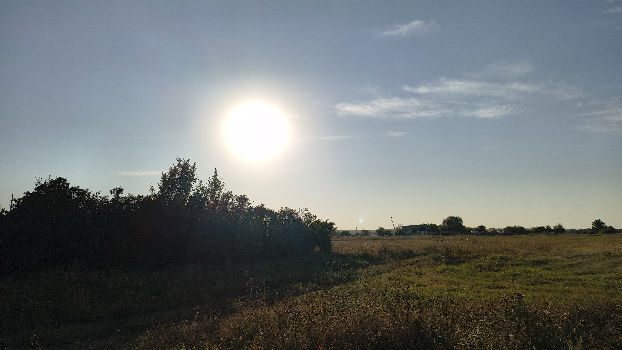 The image depicts a rural landscape under a clear blue sky with a bright sun high in the sky, casting a bright glow. In the foreground, there is a dense line of dark green trees and foliage. Beyond the trees, a wide expanse of dry, grassy fields stretches into the distance. The fields are dotted with sparse, low vegetation and appear to be agricultural land. In the far background, silhouetted against the horizon, are several buildings, some appearing to be houses or farm structures, and a few scattered trees. A power pole is visible on the left side of the frame. The overall impression is of a serene, sunlit rural scene, possibly in the morning or late afternoon, given the low angle of the sun. The location is identified as Sharapenskiy, Russia.