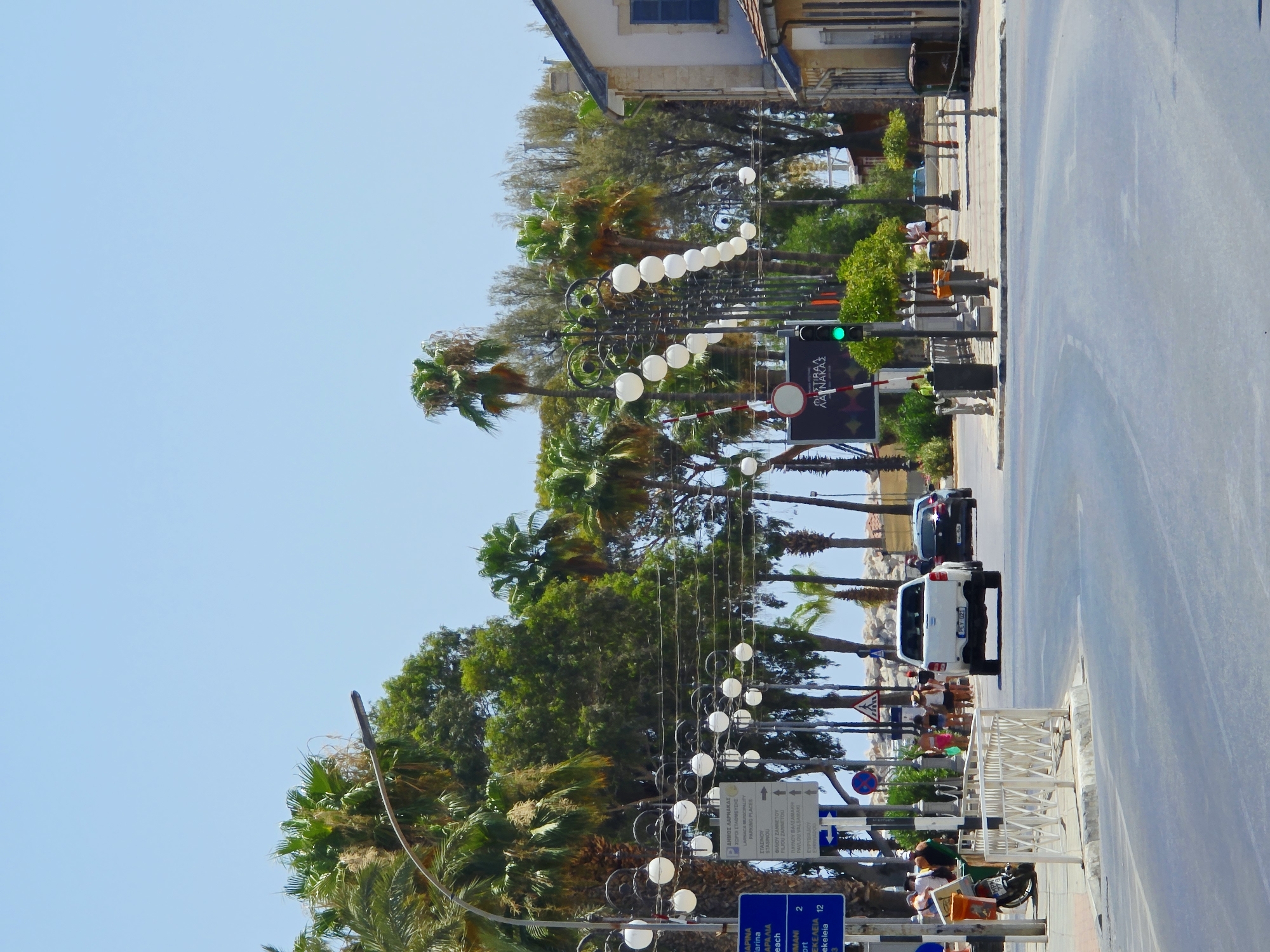 The image depicts a street scene in Larnaca, Cyprus, on a clear, sunny day. The foreground shows a paved road curving to the right, with a white pickup truck and a dark-colored car driving away from the viewer. A white railing lines the left side of the road, separating it from a sidewalk where several people are standing and walking. A white scooter is parked near the railing.
Further down the street, palm trees and other lush greenery are visible, creating a tropical atmosphere. Ornate streetlights with multiple white spherical lamps are strung along the sidewalk. A traffic signal displays a green light. Several blue and white directional signs are mounted on poles, indicating directions to various locations. A building with a light-colored facade and a dark blue shuttered window stands on the right side of the street. The sky is a clear, pale blue.