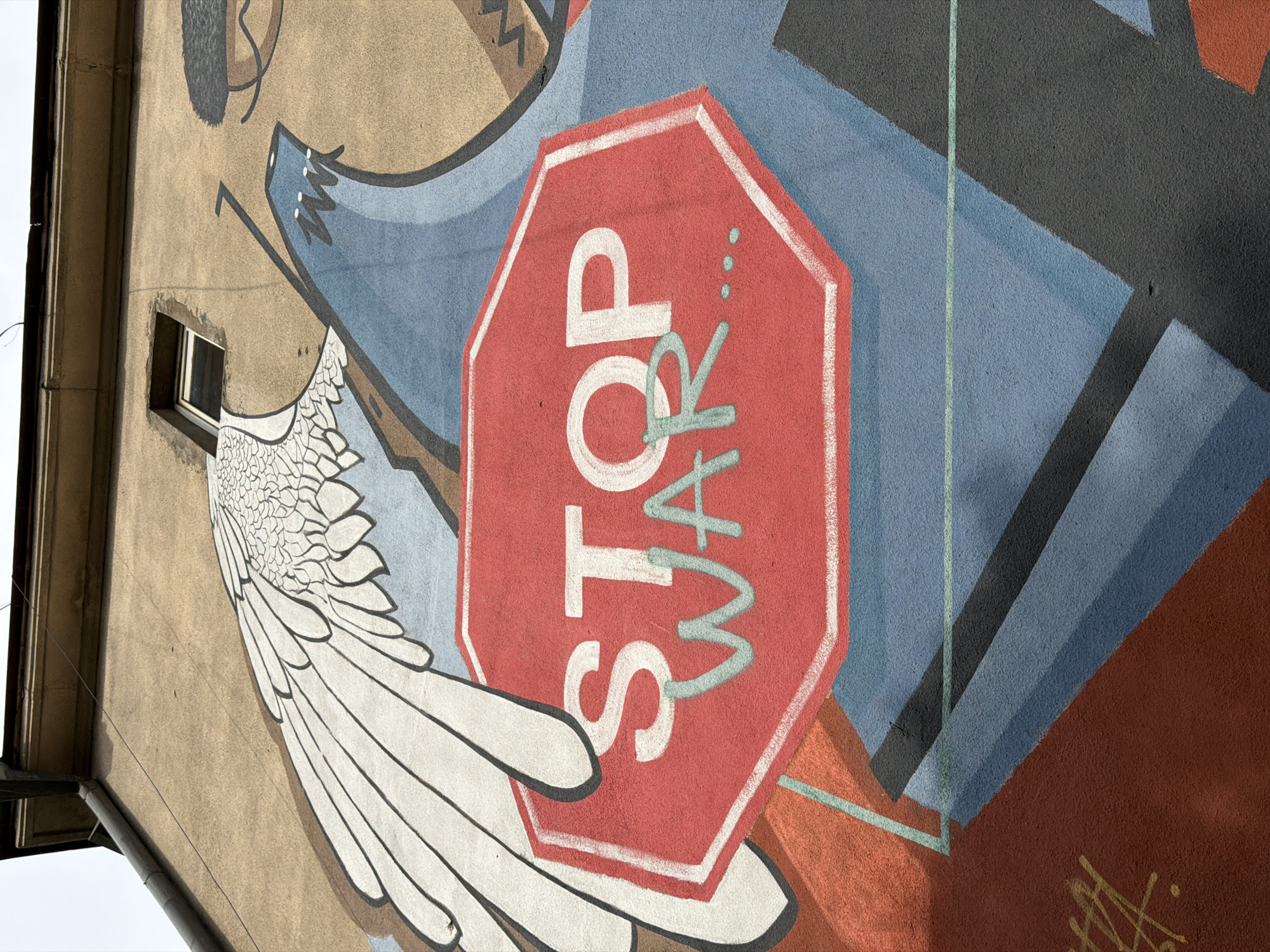 This image displays a vibrant mural painted on the side of a building, captured from a low angle looking upwards. The central and most prominent element of the mural is a large, red octagonal stop sign. Bold white capital letters spell "STOP" across the sign. Overlaid and slightly beneath the word "STOP," in a lighter blue-green hue, are the stylized letters "WAR," clearly conveying the anti-war message "STOP WAR."
To the left of the stop sign, a large, intricately detailed white wing, resembling that of an angel or a bird, extends downwards, with individual feathers distinctly outlined. Above and to the left of this wing, a small, dark-framed rectangular window is set into the building's textured, beige-brown wall. The rest of the mural is composed of abstract, geometric shapes in various shades of blue, gray, and brown, creating a dynamic backdrop.
The building wall itself appears to be made of stucco or painted concrete, showing some signs of age and texture. In the bottom right corner of the mural, some abstract, graffiti-like markings or signatures in yellow and black are visible. The bright, even lighting suggests it is daytime. The scene evokes a strong anti-war sentiment through its direct message and symbolic imagery, such as the wing, which might represent peace or hope. The location context is Tbilisi, Georgia.