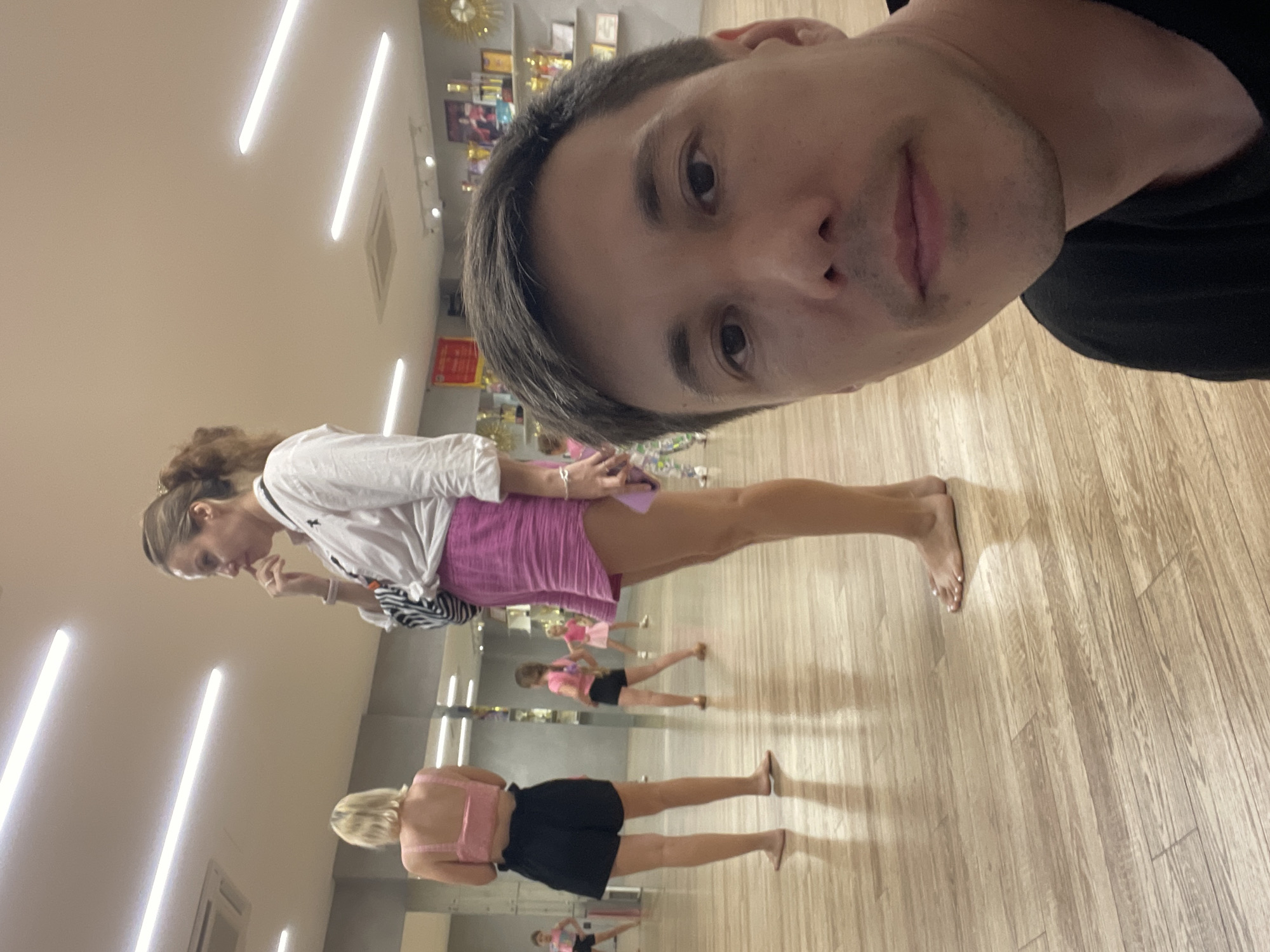 The image shows a group of people in what appears to be a dance studio or a similar indoor space with a polished wooden floor and bright fluorescent lighting. The perspective suggests a selfie taken by a man in the foreground, his face close to the camera. He is wearing a black t-shirt.
In the mid-ground, a woman with brown hair tied up is looking downwards, touching her finger to her mouth. She is wearing a white long-sleeved shirt and a pink skirt. Behind her, other women are visible, also in athletic or dance attire, some appearing to be in formation, suggesting a class or practice session. One woman with blonde hair is wearing a pink top and black shorts, and another in the background has on a pink top and black shorts as well. Mirrors on the wall reflect the scene, showing more individuals in the studio.
The overall mood seems casual and active, possibly indicative of a dance lesson. The lighting is bright, suggesting it could be daytime or that the studio is well-lit regardless of the time of day. The setting in Thủ Đức, Vietnam, provides a geographical context. There are no obvious signs of text in the image.