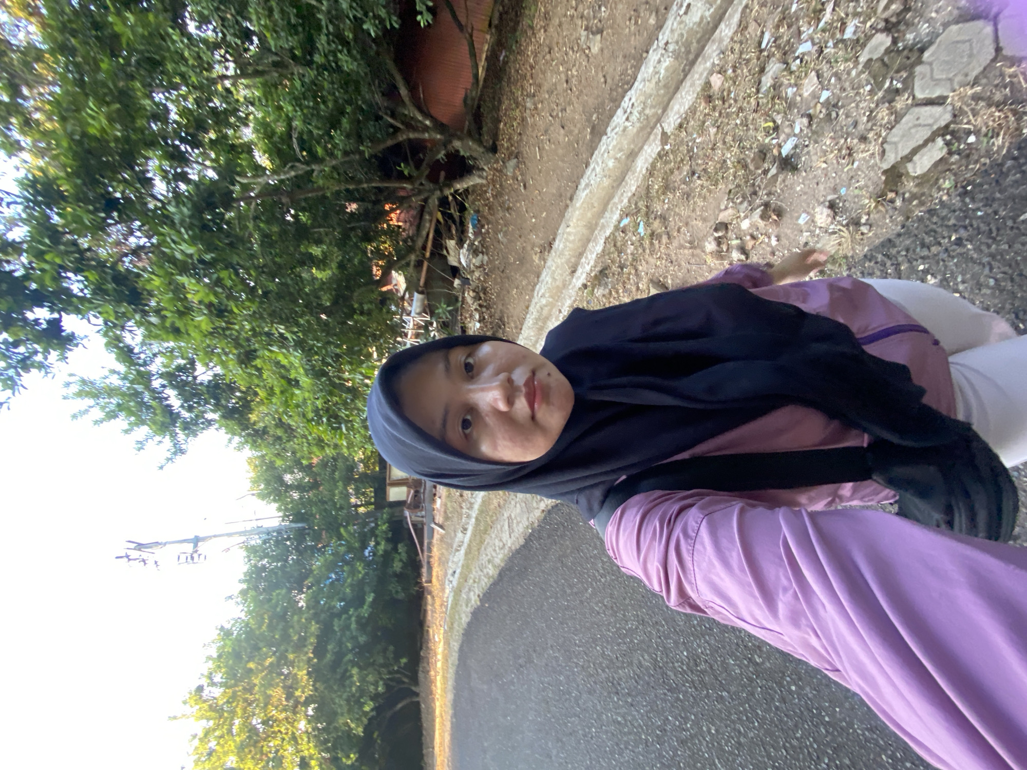 A young Indonesian woman wearing a hijab and a light purple jacket is taking a selfie outdoors. She is standing on a paved pathway with trees and vegetation in the background. A cell tower can be seen in the distance. The image appears to be taken in Banjarbaru, Indonesia, given the context provided.