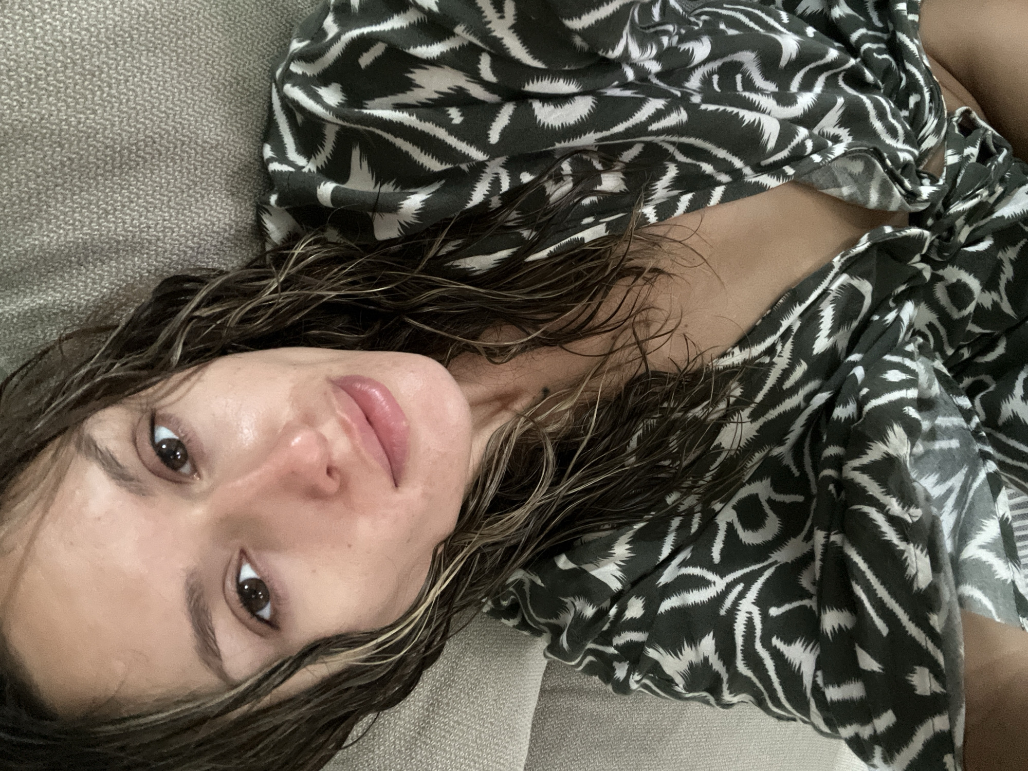 A woman with wet, dark brown hair is lying on a textured, grey couch. She is wearing a black and white patterned robe. The woman's face is turned towards the camera, and her eyes are open. Her lips are slightly parted. The lighting is soft and natural. There are no other people or objects visible in the image. The background is the grey couch. The location is a private residence in the city of Choeng Thale, Thailand.