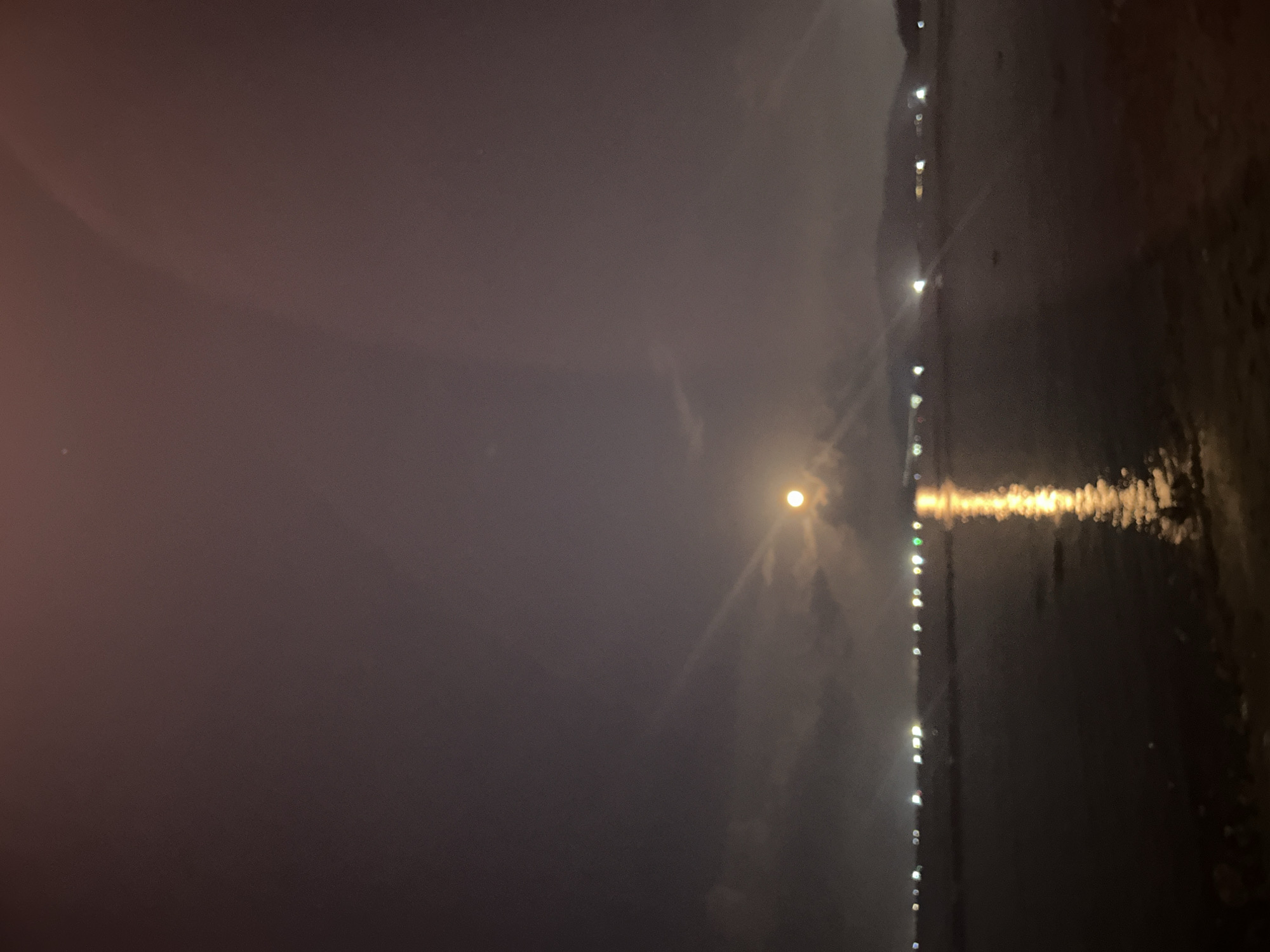 The image captures a nighttime scene in Bac Nha Trang Ward, Vietnam. A body of water reflects the light of street lamps along the coast, as well as the light of the moon. The sky is mostly obscured by clouds or haze.