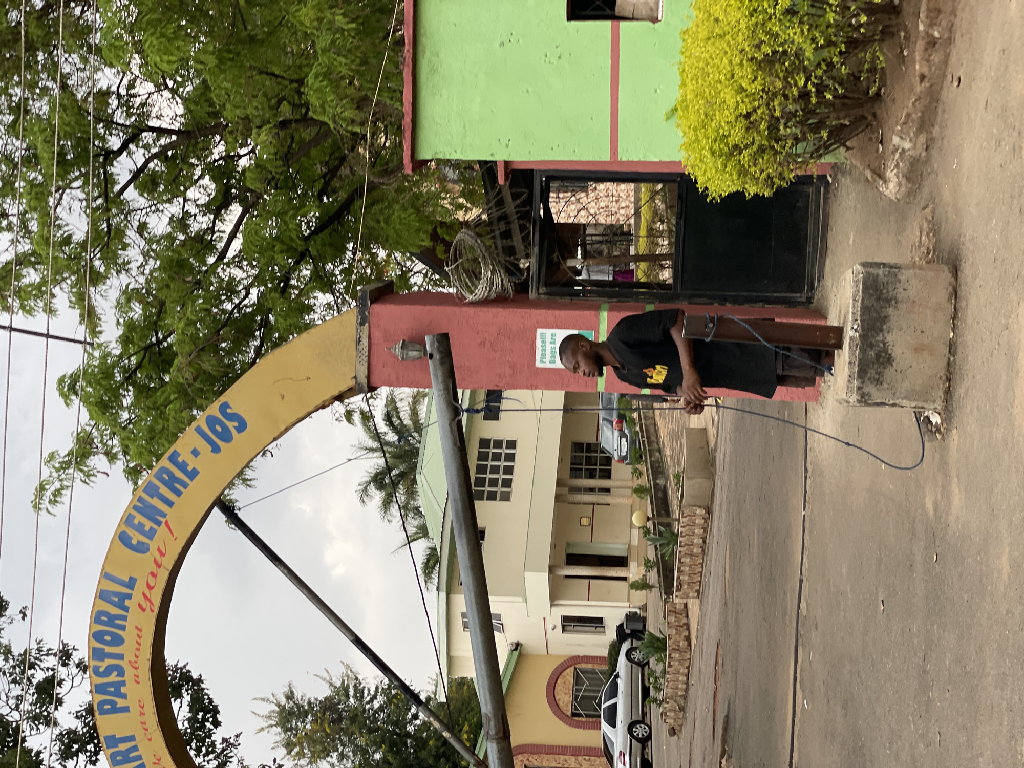 The image shows an outdoor scene in what appears to be Jos, Nigeria. A sign reading "ART PASTORAL CENTRE - JOS" in blue letters on a yellow archway is visible. The entrance is flanked by a red and green painted building. A man in a black t-shirt is standing near a concrete structure and is holding a blue cable. In the background, a two-story building with white walls and multiple windows is visible, along with parked cars. Trees are scattered throughout the scene, and the sky is overcast.