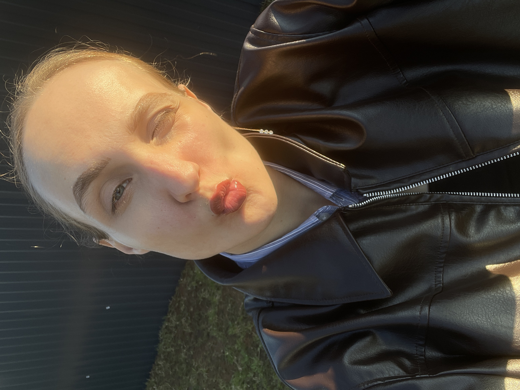 A young woman with light hair is depicted in a close-up, rotated selfie, making a playful expression with pursed, glossy lips. She is wearing a black faux leather jacket over a blue and white striped shirt. A delicate necklace with three small, light-colored beads or stones is visible around her neck.
The photograph is taken outdoors during the golden hour, likely late afternoon, as warm sunlight strongly illuminates her face and the left side of her body, creating distinct highlights and shadows. Her skin appears fair, and her eyebrows are well-defined. Her left eye, partially visible, appears to be a light color.
The background consists of a dark, corrugated fence or wall, suggesting an outdoor, possibly urban or suburban setting. Below, patches of green foliage or grass are visible, adding to the outdoor ambiance. The overall mood of the image is casual and lighthearted. There is no visible text in the image.