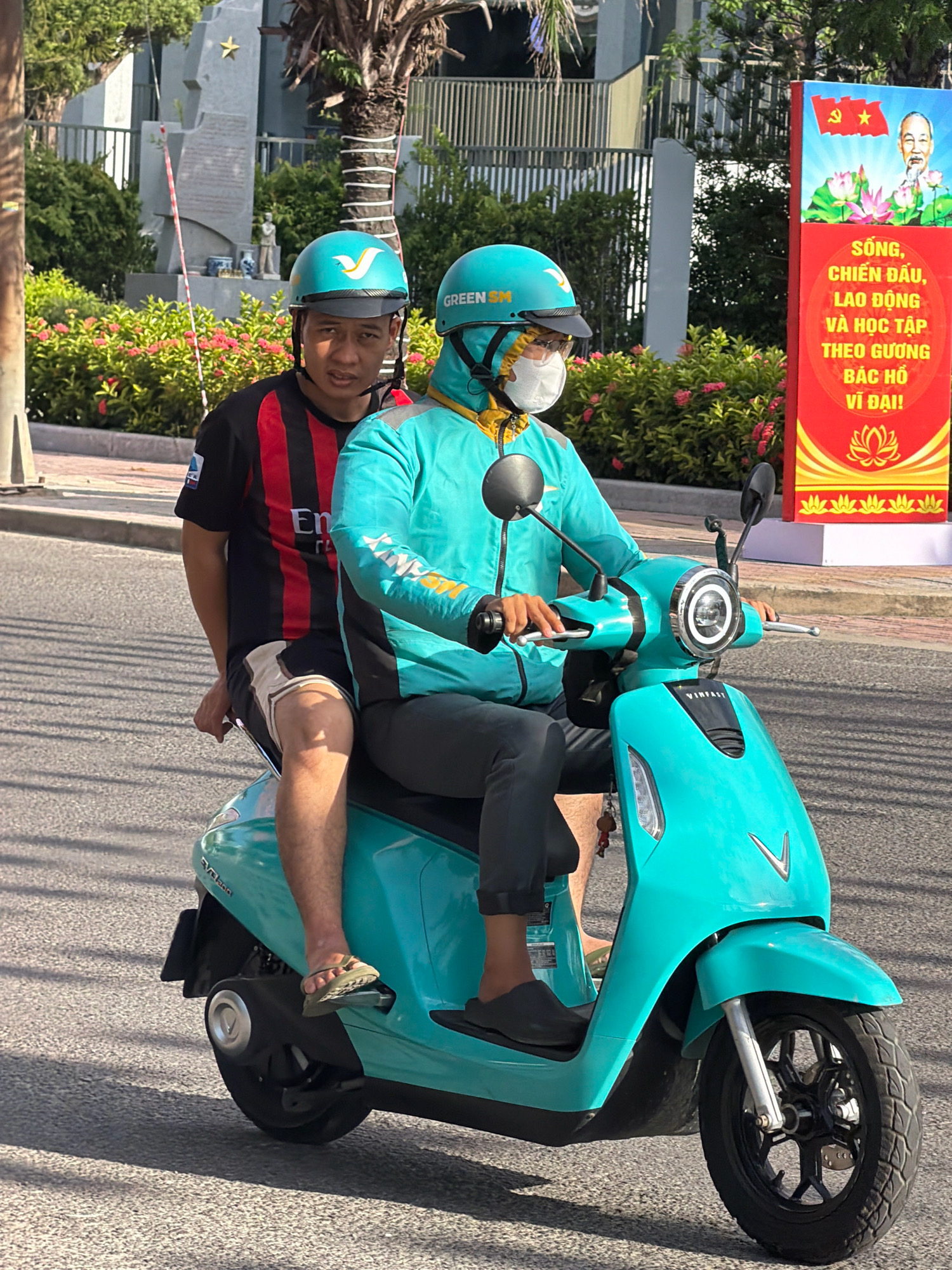 Two men are riding a teal electric scooter in Nha Trang, Vietnam. The man in the front is wearing a turquoise jacket, a white face mask, and a teal helmet. He is holding the handlebars and appears to be looking forward. The man in the back is wearing a black and red striped t-shirt and tan shorts with black stripes. He is wearing a teal helmet and has his arms around the front rider. His left leg is visible, wearing a tan sandal. The scooter has a prominent circular headlight. In the background, there is a monument with a star on top, surrounded by green bushes and pink flowers. To the right, a red banner displays an image of Ho Chi Minh and Vietnamese text, along with lotus flowers. The scooter is on a paved road with a sidewalk to the left. Sunlight is casting shadows on the road.