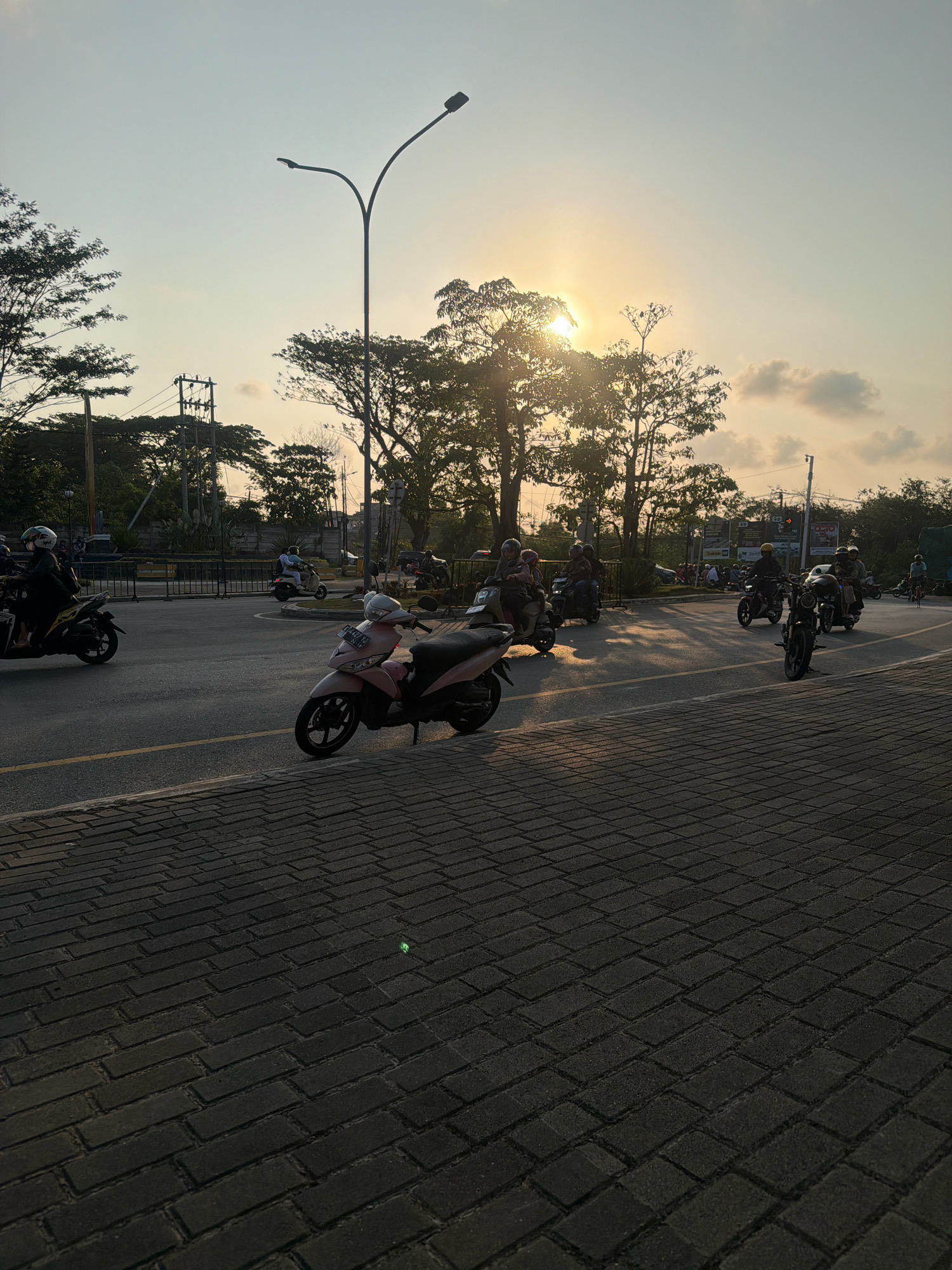 The image depicts a street scene at sunset in Balikpapan, Indonesia. The sun is low in the sky, casting a warm glow and long shadows. Several motorcycles, most with riders wearing helmets, are present on the road. One pink scooter is parked in the foreground on a brick-paved sidewalk. In the background, there are trees, power lines, and streetlights. The sky is a pale yellow and orange, with some scattered clouds. The overall impression is one of daily life in a tropical city during the late afternoon.