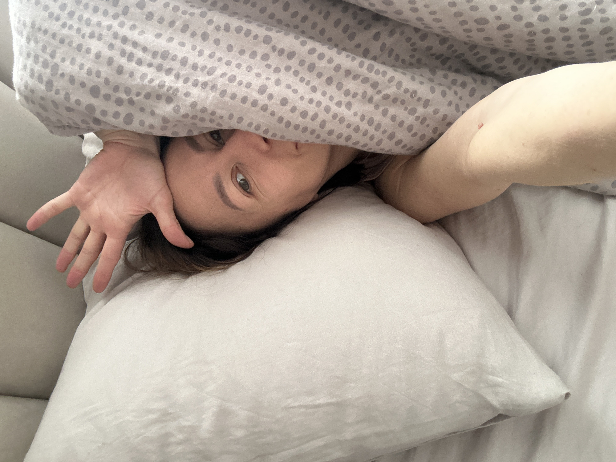 A woman lies in bed with a light gray polka dot patterned blanket pulled partly over her face. She is resting her head on a white pillow. Her left hand is extended, palm facing the camera. Her right arm is also visible. The setting appears to be an indoor bedroom. There is no information to suggest the location is Votkinsk, Russia.
