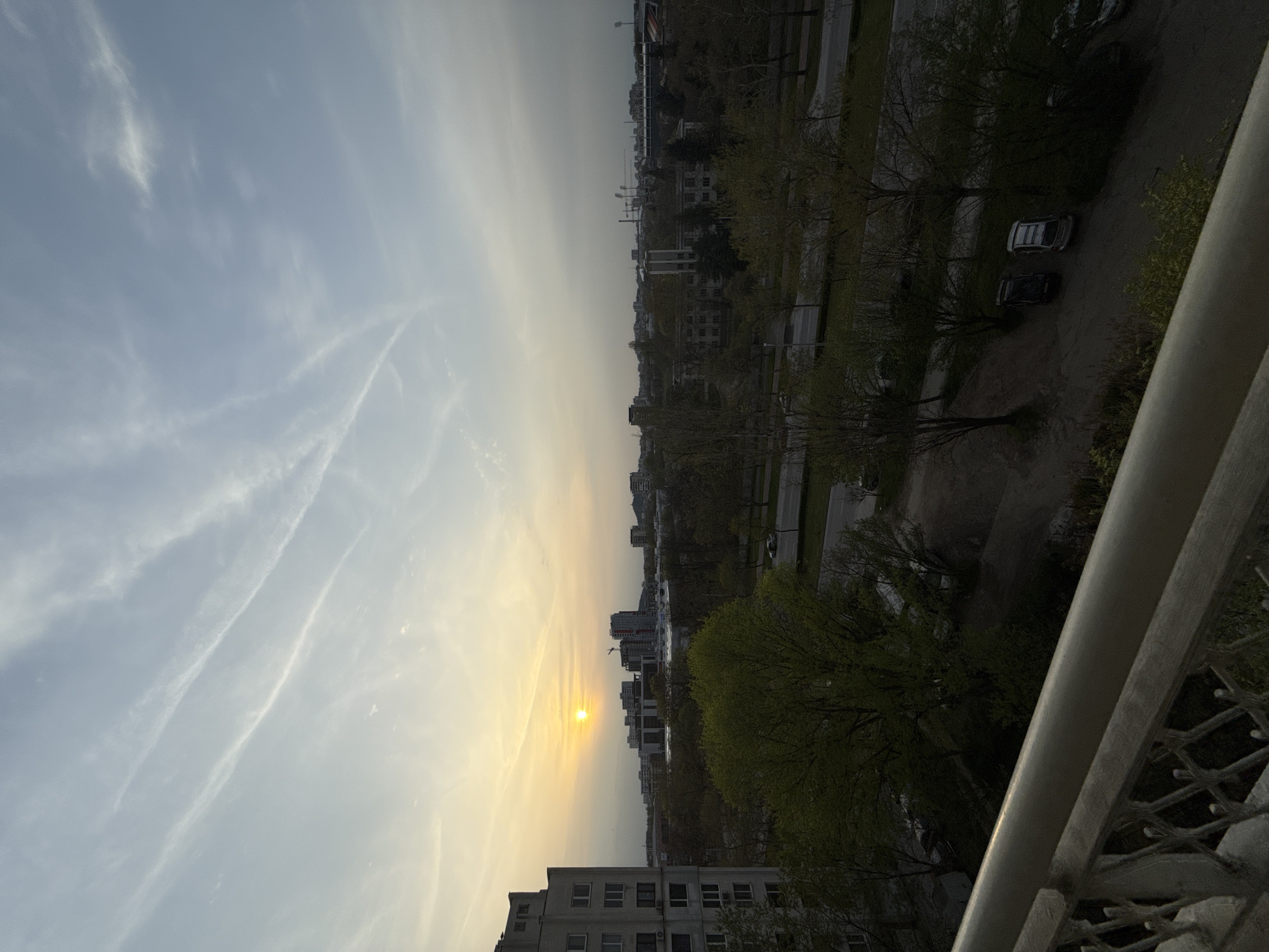The image depicts a sunset view over an urban landscape, likely taken from a high vantage point like a balcony. The sun is low on the horizon, creating a yellow and orange glow in the sky. Cirrus clouds are visible. Below the horizon, city buildings and green trees stretch into the distance. A couple of cars are visible in the lower right. The railing of the balcony is also visible, which provides a frame for the scene. Based on the buildings, the image may be in Пловдив, Bulgaria. A stadium is barely visible in the distance.