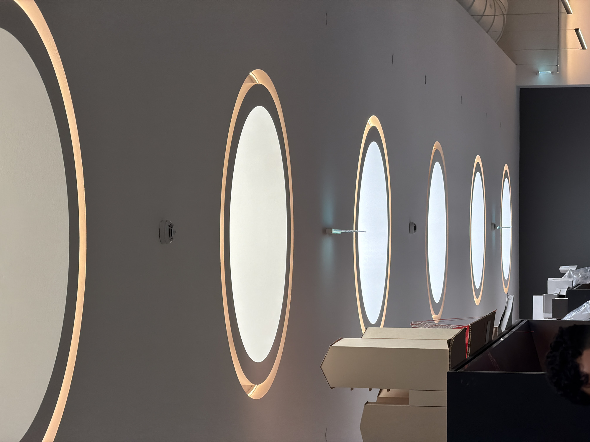 The image shows a gray wall with several oval-shaped light fixtures arranged in a row, diminishing in size from left to right, suggesting a receding perspective. The wall is gray and has a fire alarm, with what appears to be a thin metal rod that is attached to the wall. In the foreground, cardboard boxes and a dark-colored cabinet partially obscure the view. Based on the style and ambiance, this interior design is most likely located in Milan, Italy. There are no people visible. The scene appears to be an interior of a building, possibly a store, gallery, or office space. The oval light fixtures are the focal point, with a bright, inner oval surrounded by a golden-colored ring of light.